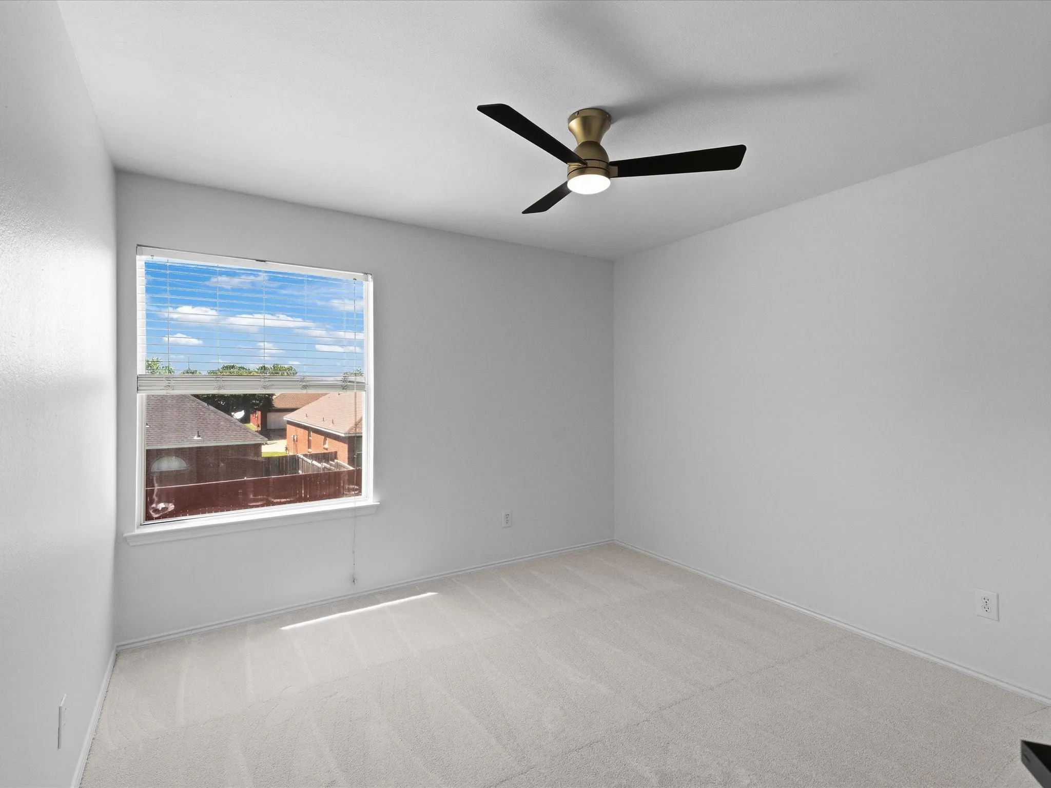 Spacious room featuring light-colored carpeting, a window with blinds, and a contemporary ceiling fan with a brass-finish base and black blades