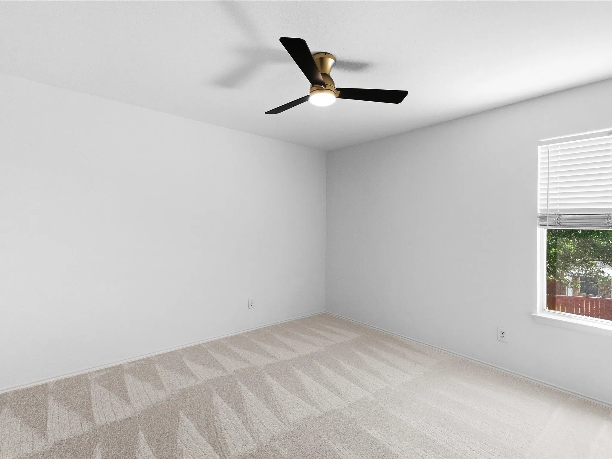 Neutral-toned room featuring a window with white blinds, a ceiling fan with black blades and a brass-finish fixture, and light-colored patterned carpeting