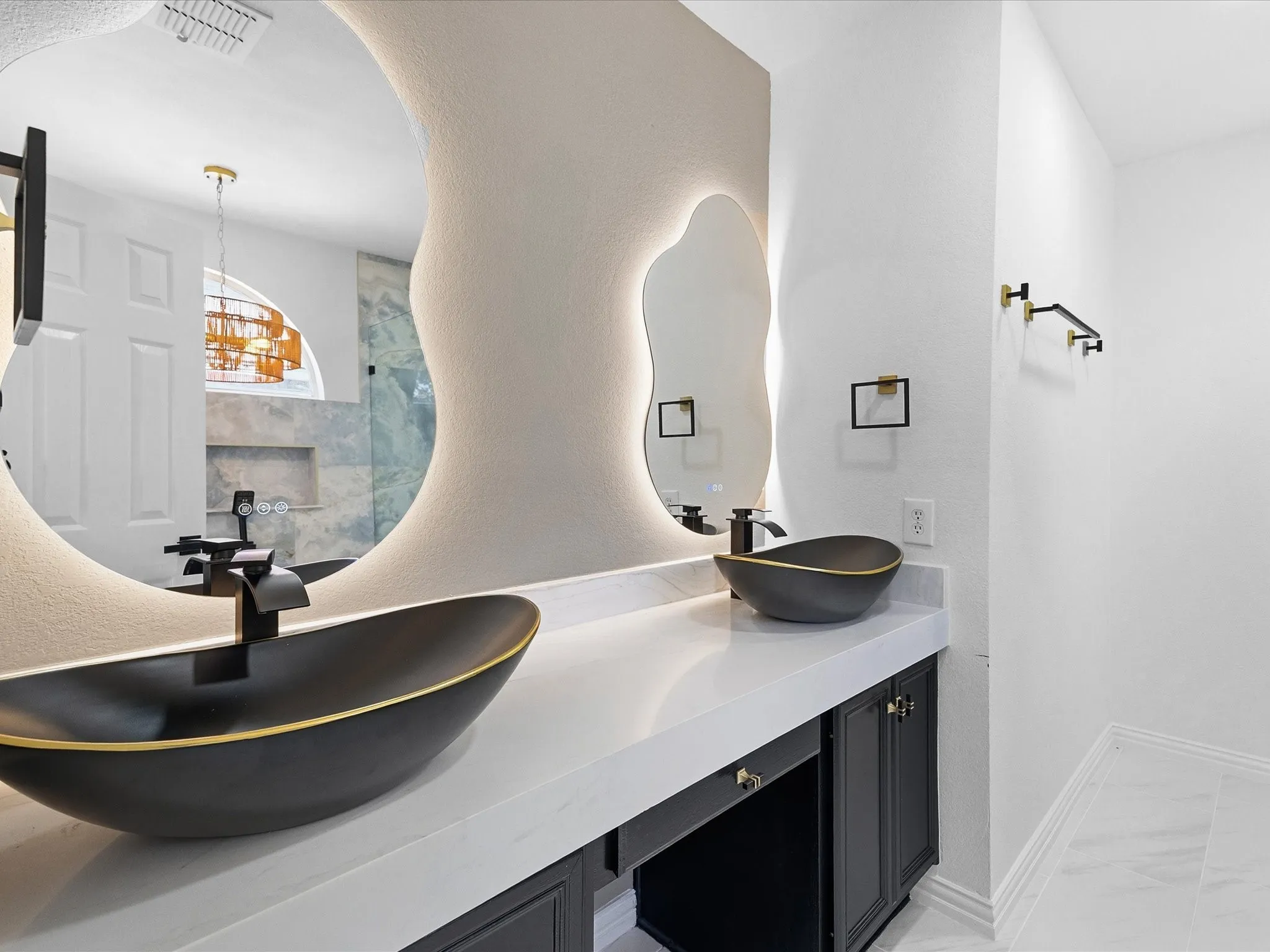 Dual vanity featuring black vessel sinks with gold accents, back-lit organic shaped mirrors, white countertop, and dark cabinetry