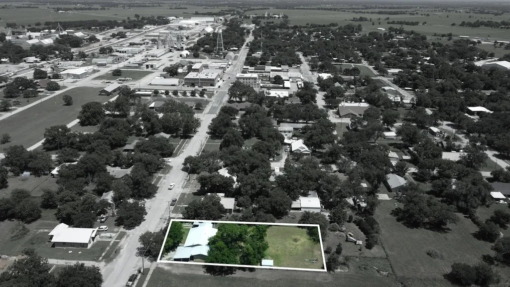 Aerial perspective showcasing a property with a tree-lined perimeter, a primary structure with a light-colored roof, and an expansive grassy area