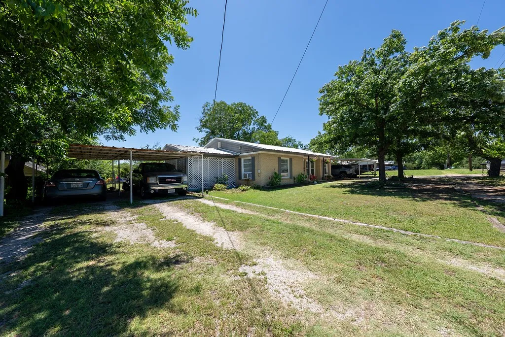 Expansive front yard featuring a multi-car carport, mature shade trees, a gravel driveway, and a brick exterior with white lattice detailing