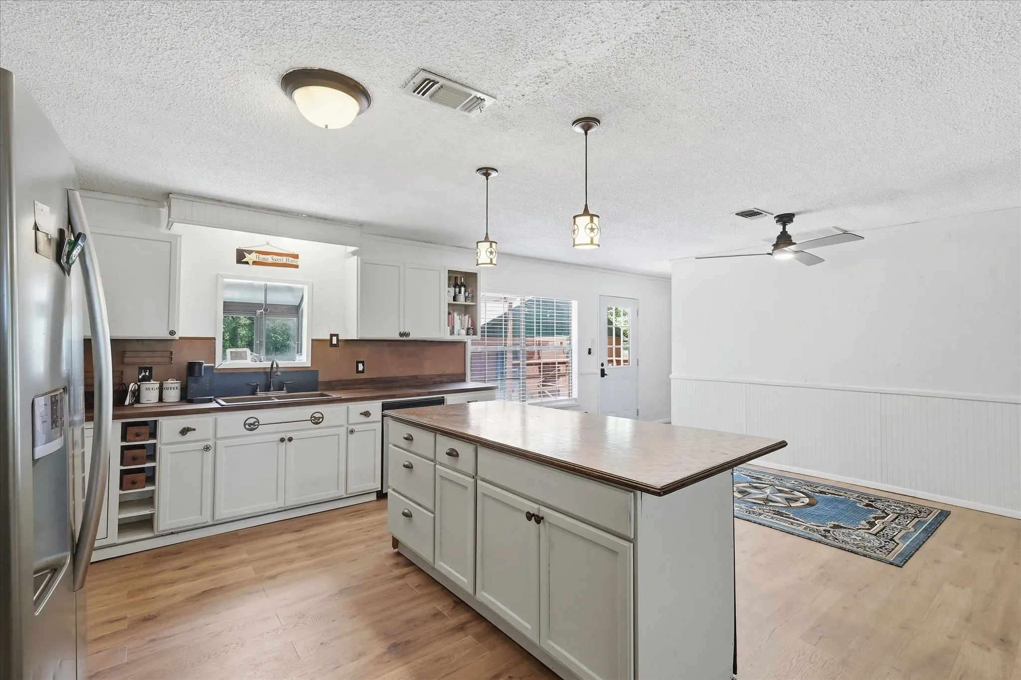 Expansive kitchen featuring a central island with countertop, white cabinetry with dark hardware, wood-finish flooring, and a stainless steel refrigerator