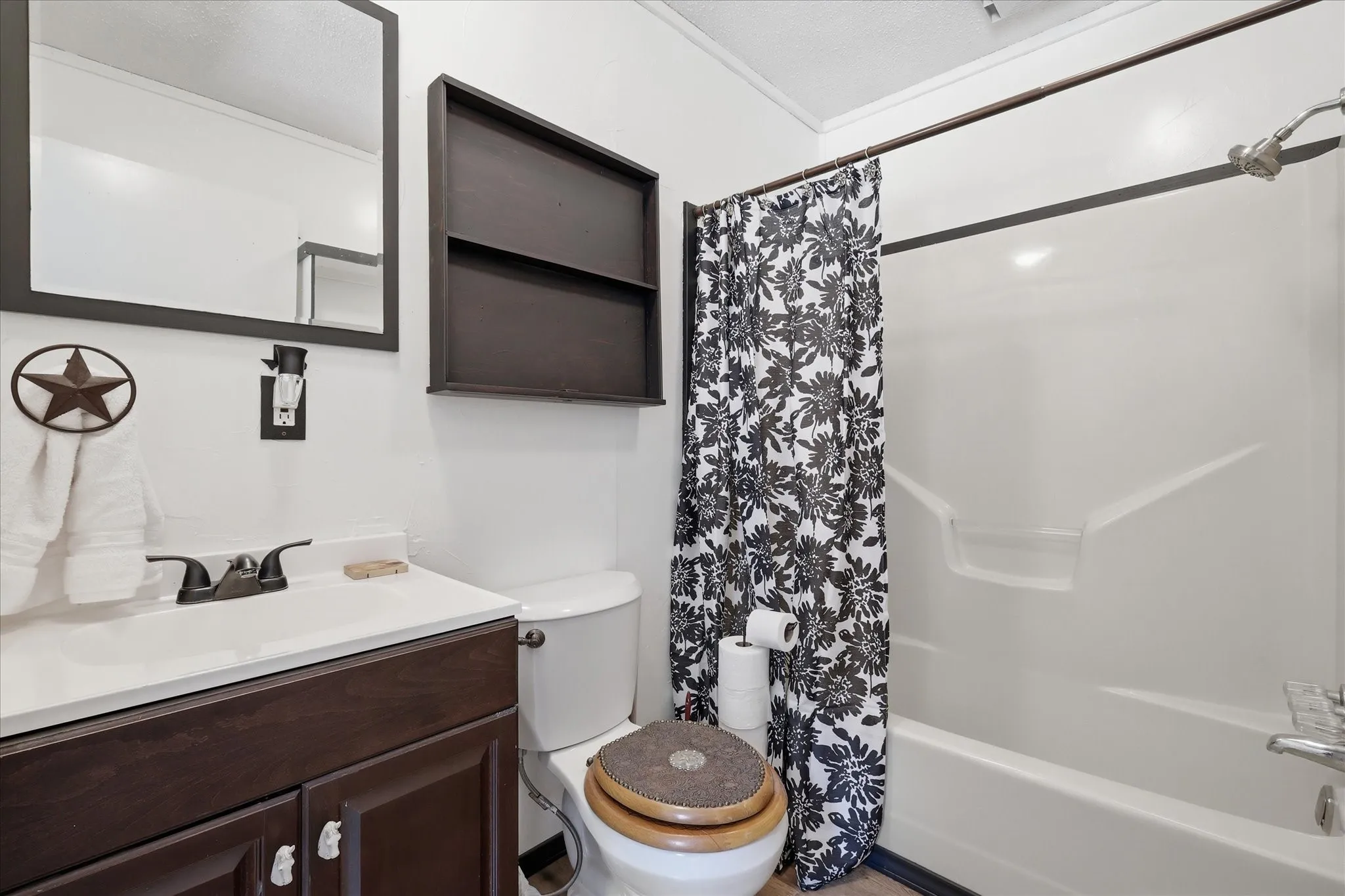 Bathroom featuring a white vanity with a dark wood-finish cabinet and a bronze-finish faucet, a white toilet with a wood-finish seat, a white tub and shower combination, a dark wood-finish framed mirror, and a dark wood-finish wall shelf