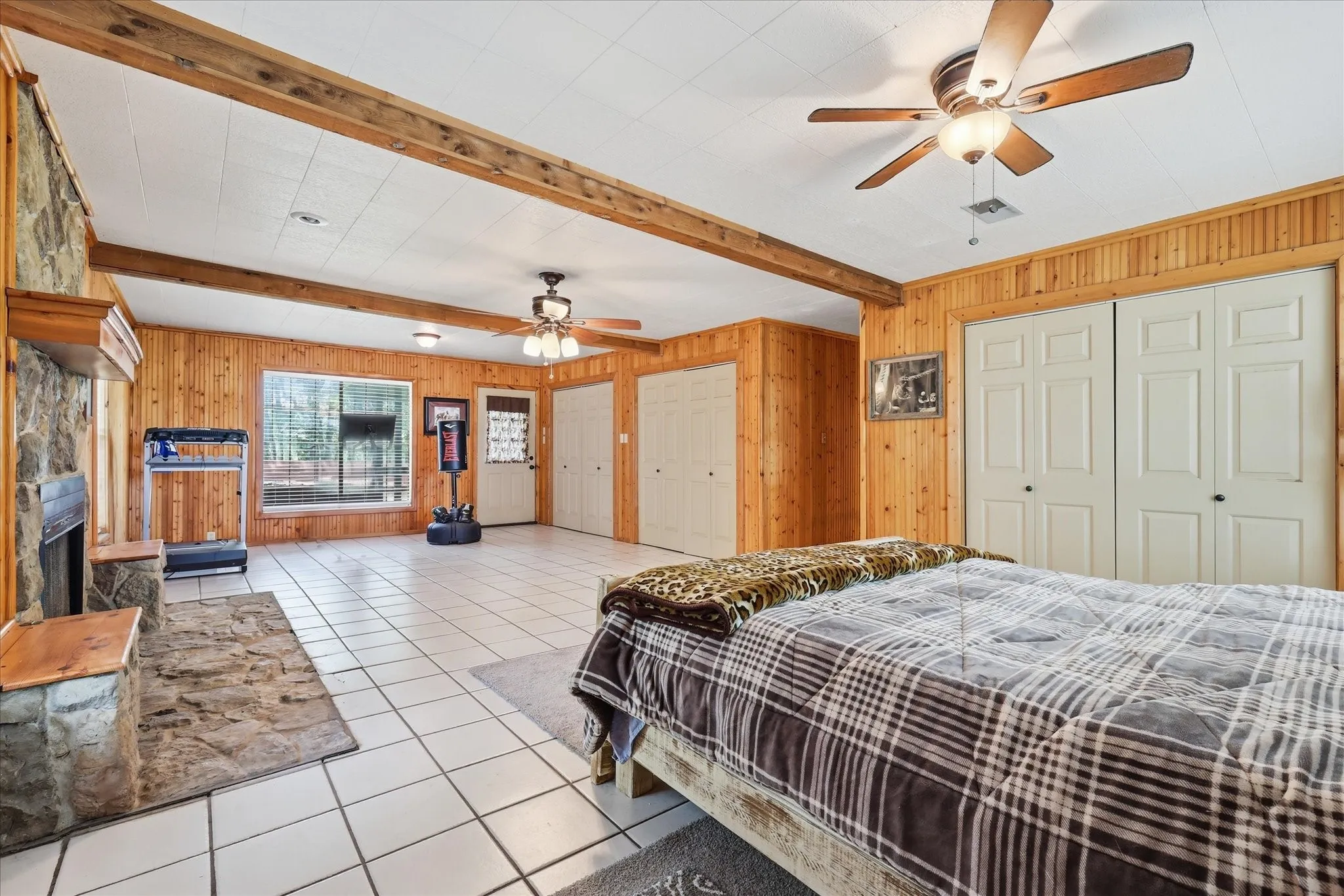 Spacious room featuring wood paneling, exposed ceiling beams, a stone fireplace with a wood mantle, white tile flooring, and multiple ceiling fans