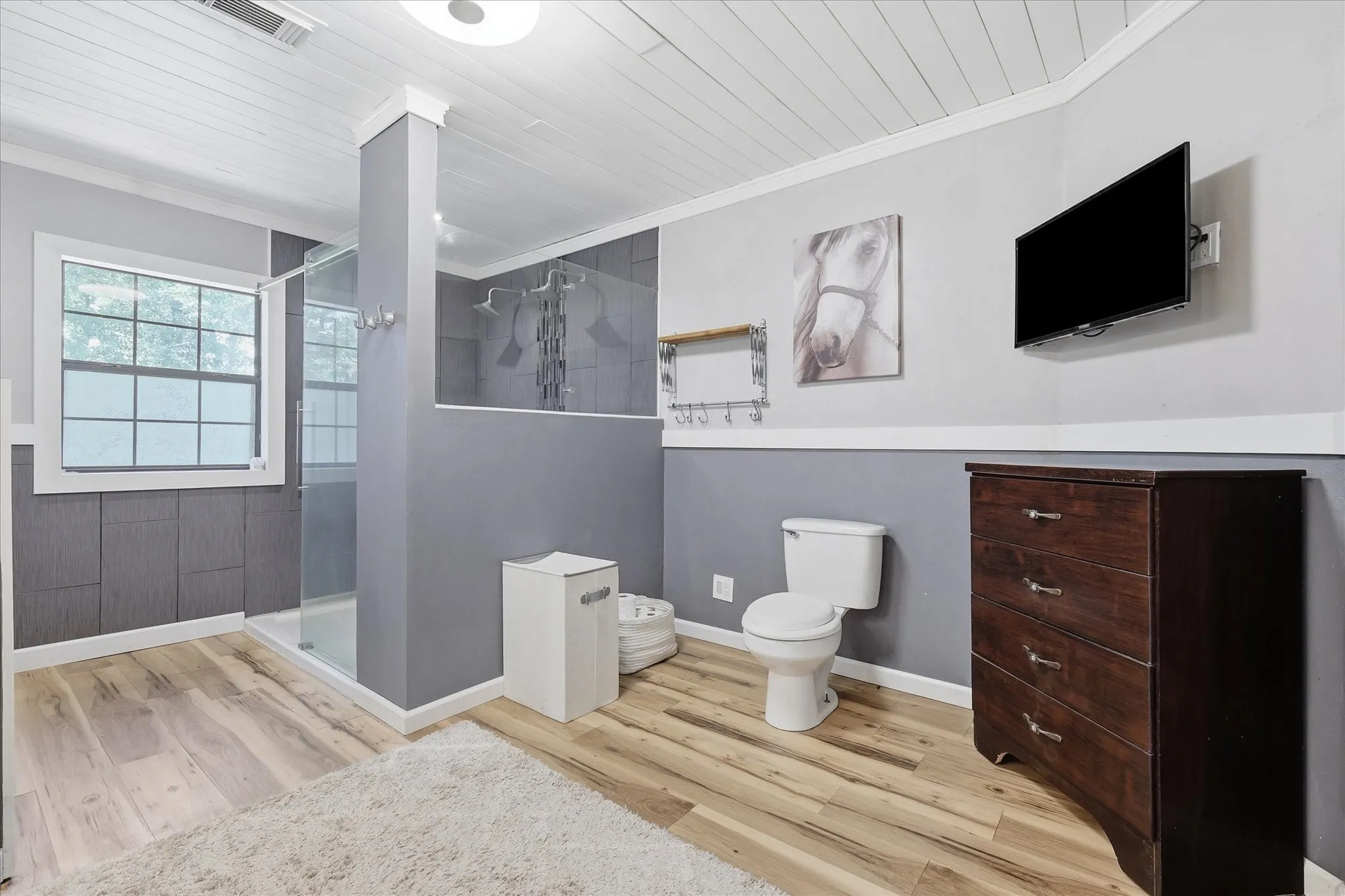 Bathroom featuring wood-finish flooring, a glass-enclosed shower with multiple showerheads, a window, and paneled walls