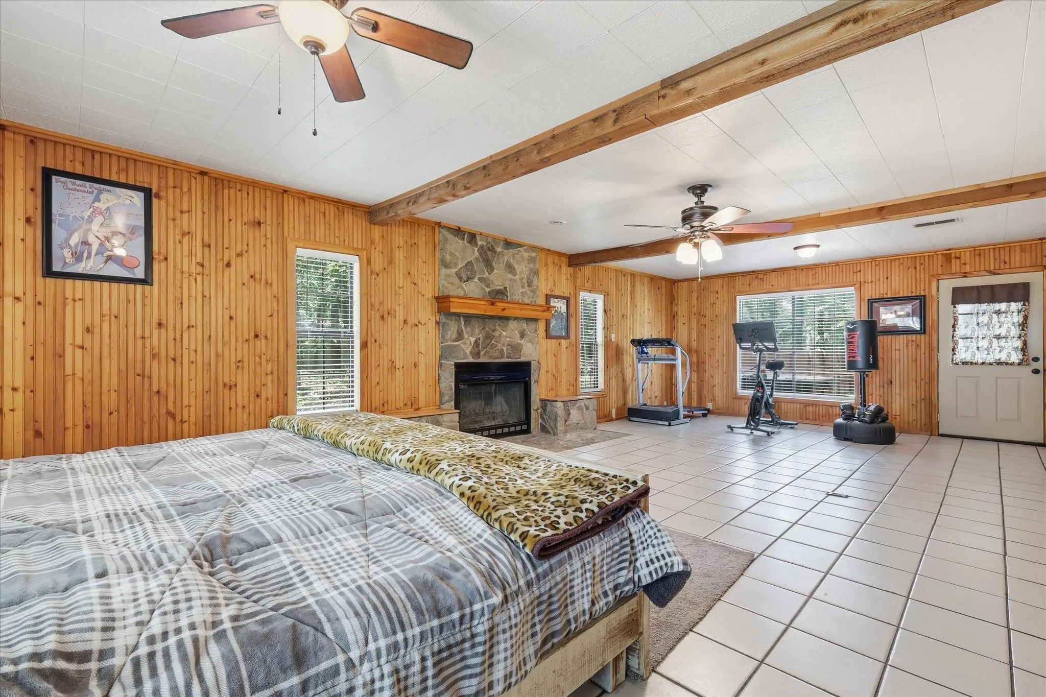 Spacious room featuring wood paneling, exposed ceiling beams, a stone fireplace with a wood mantel, tile flooring, and multiple windows