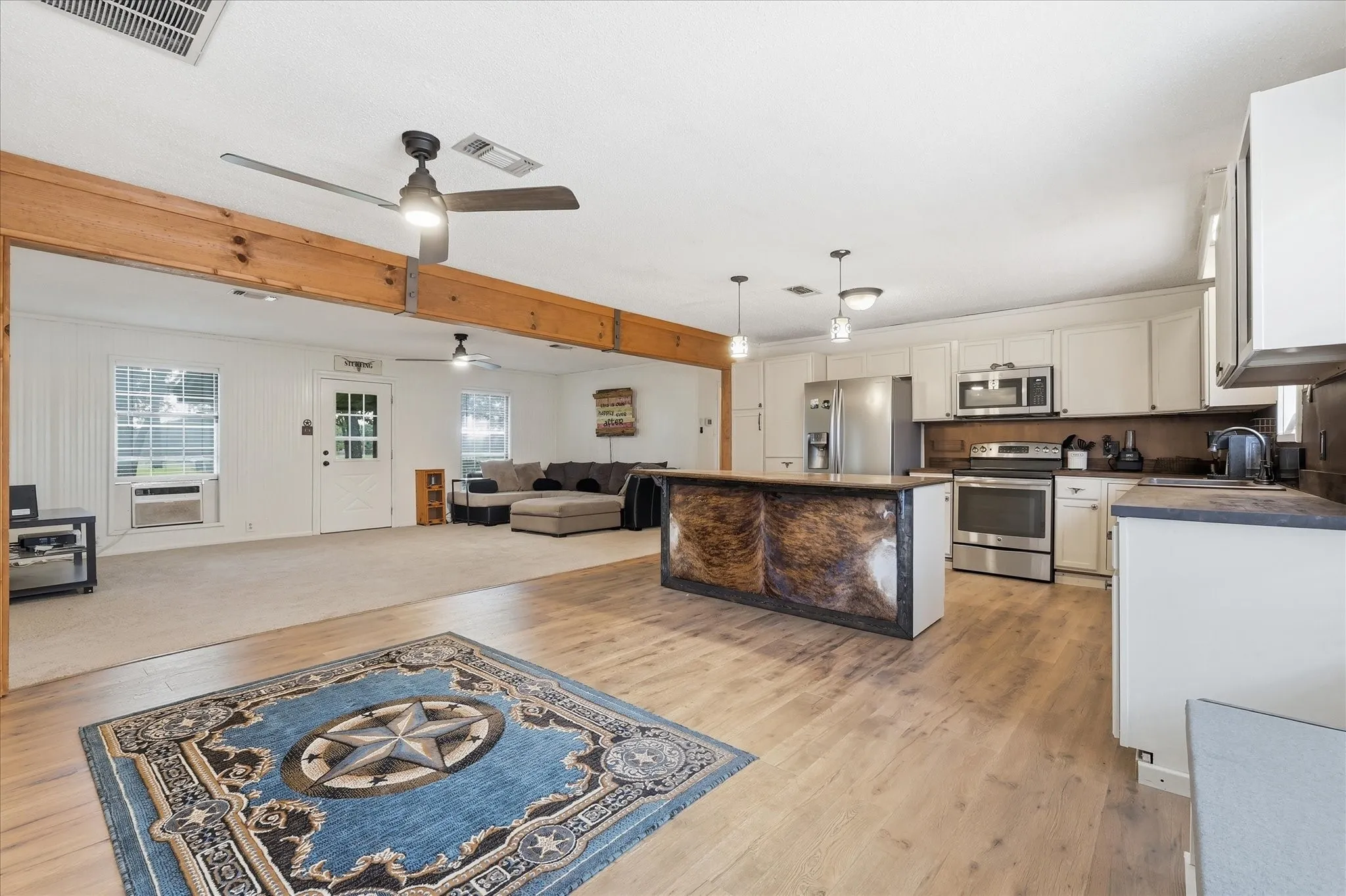 Open-concept kitchen and living area featuring wood-finish flooring, a central kitchen island with pendant lighting, stainless steel appliances, and white cabinetry