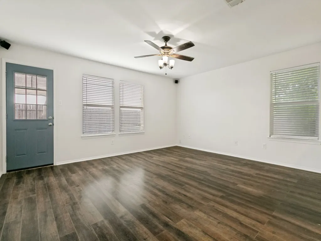 Spacious living area featuring wood-finish flooring, white walls, and multiple windows with blinds