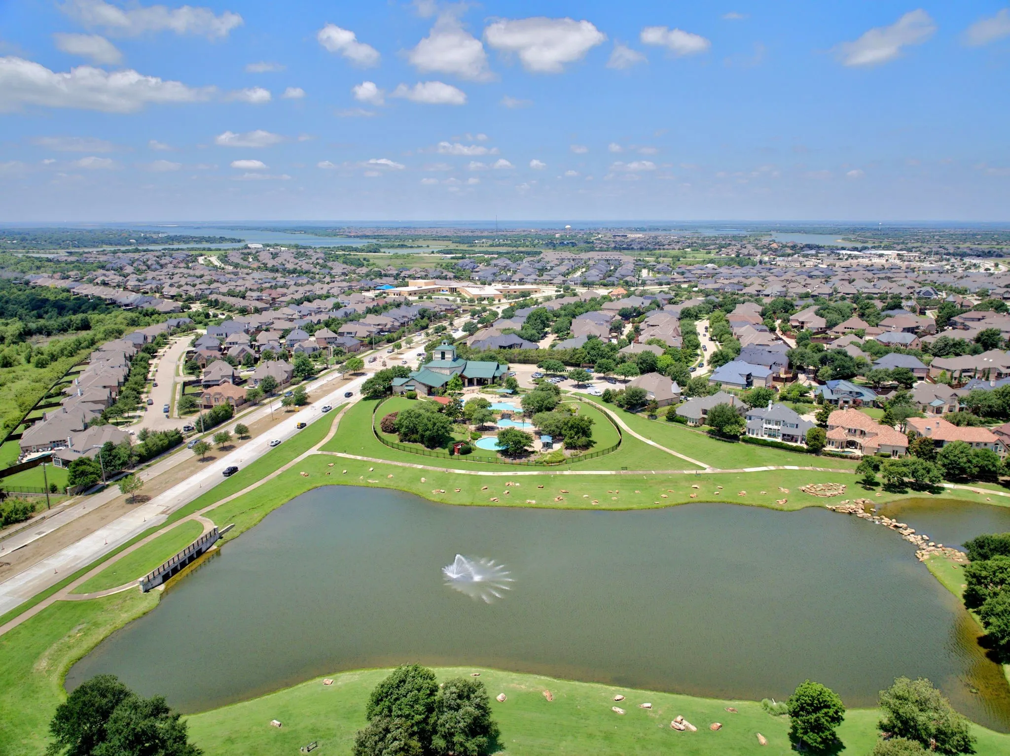 Expansive aerial showcasing a residential community with numerous single-family homes, a large central pond with a fountain, and green spaces