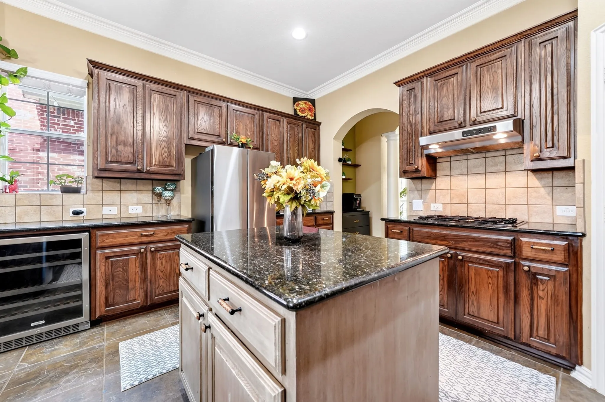 Kitchen featuring a central island with a dark speckled countertop, rich wood-finish cabinetry, stainless steel appliances, a built-in wine refrigerator, and a tile backsplash