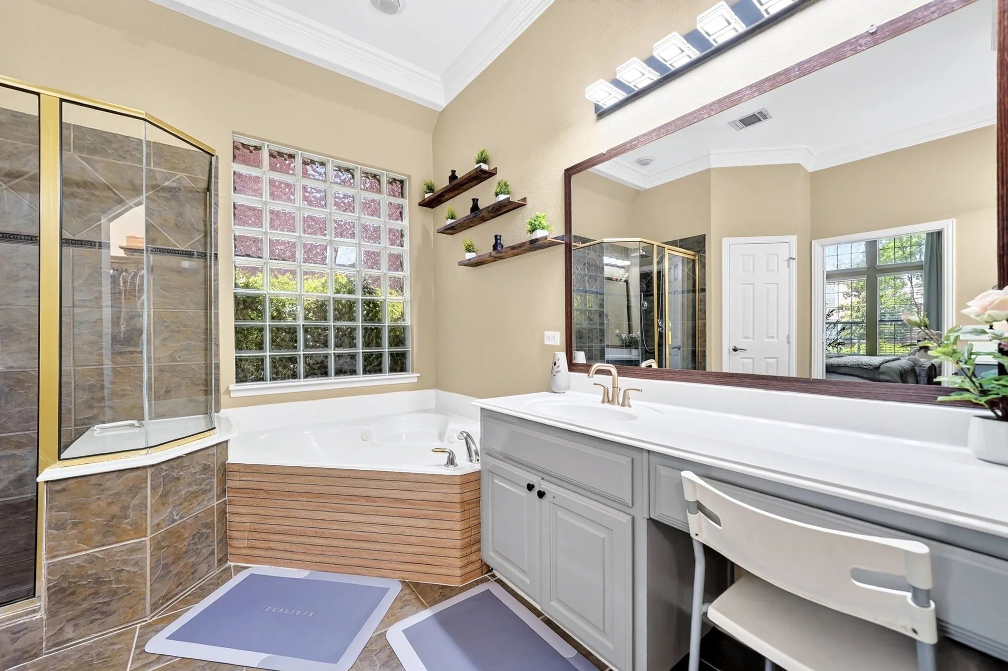 Bathroom featuring a corner jetted tub with a wood-finish surround, a glass block window, a glass-enclosed shower with gold-tone framing, a double vanity with light gray cabinetry, and a large wall-mounted mirror