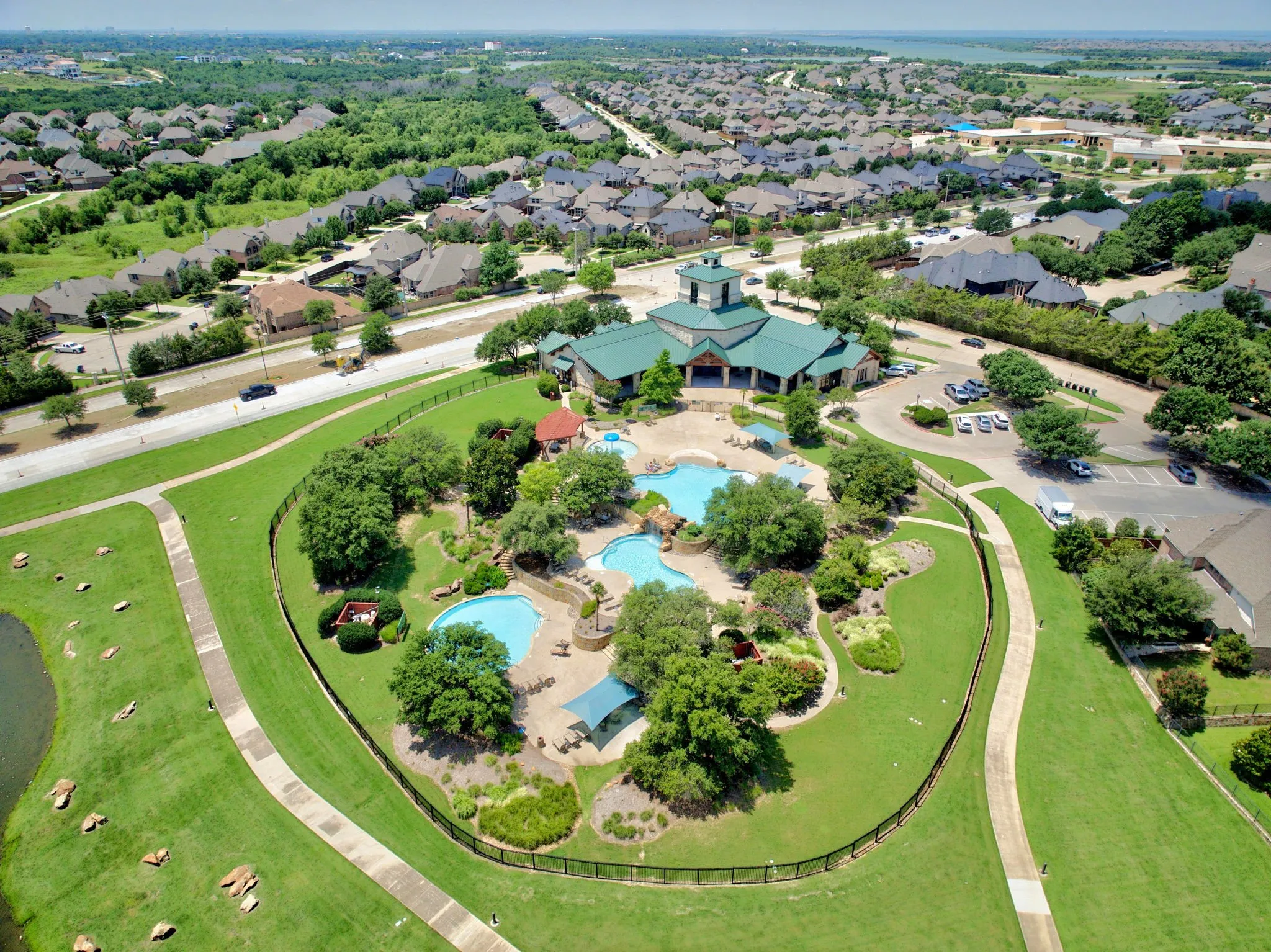 Community amenities featuring multiple swimming pools with rock features, a central clubhouse building with a green roof, and extensive green space with walking paths