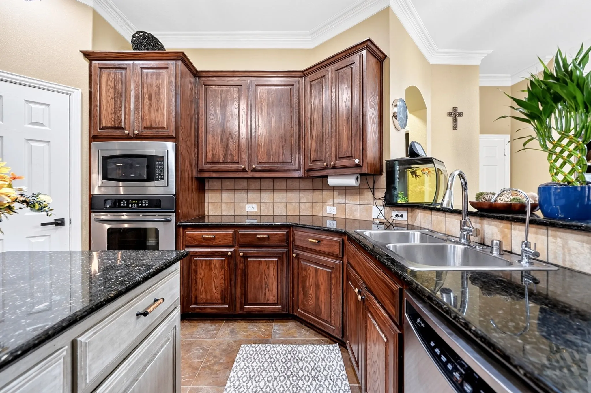 Kitchen featuring dark wood cabinetry, stainless steel appliances including a built-in oven and microwave, dark speckled countertops, a tile backsplash, and a double basin stainless steel sink