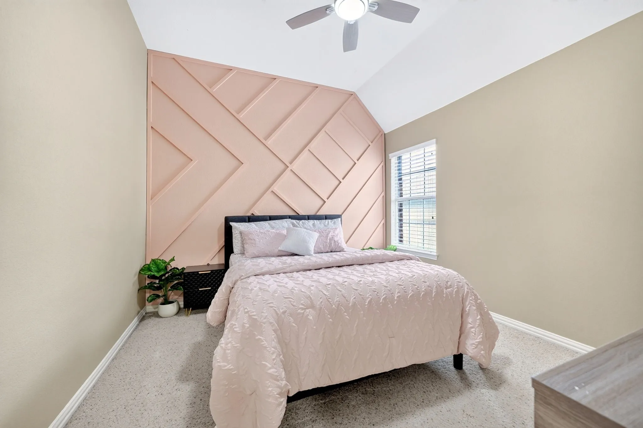 Vaulted ceiling with a ceiling fan, geometric accent wall, natural light from a window with blinds, carpet flooring, and neutral wall paint