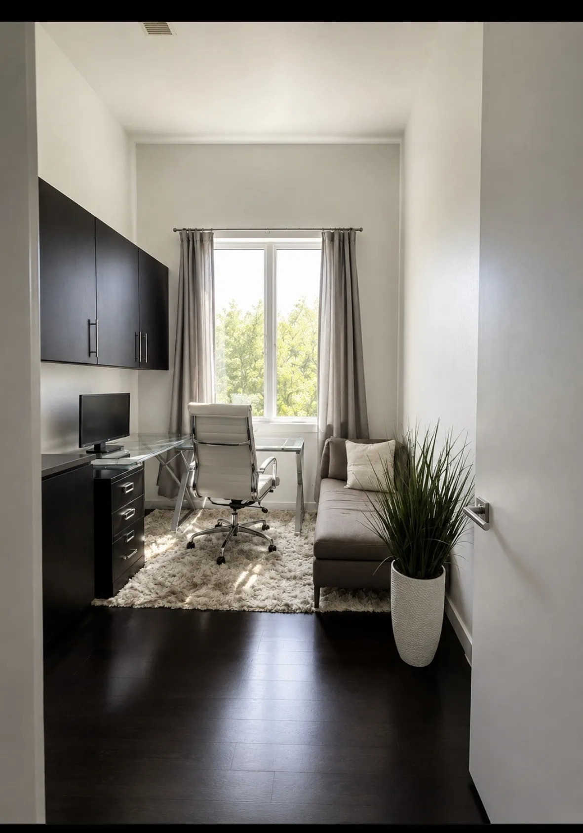 Modern home office featuring dark wood-finish flooring, wall-mounted flat-screen display, built-in desk cabinetry with drawer storage, large multi-pane window with natural light, and contemporary gray upholstered task seating