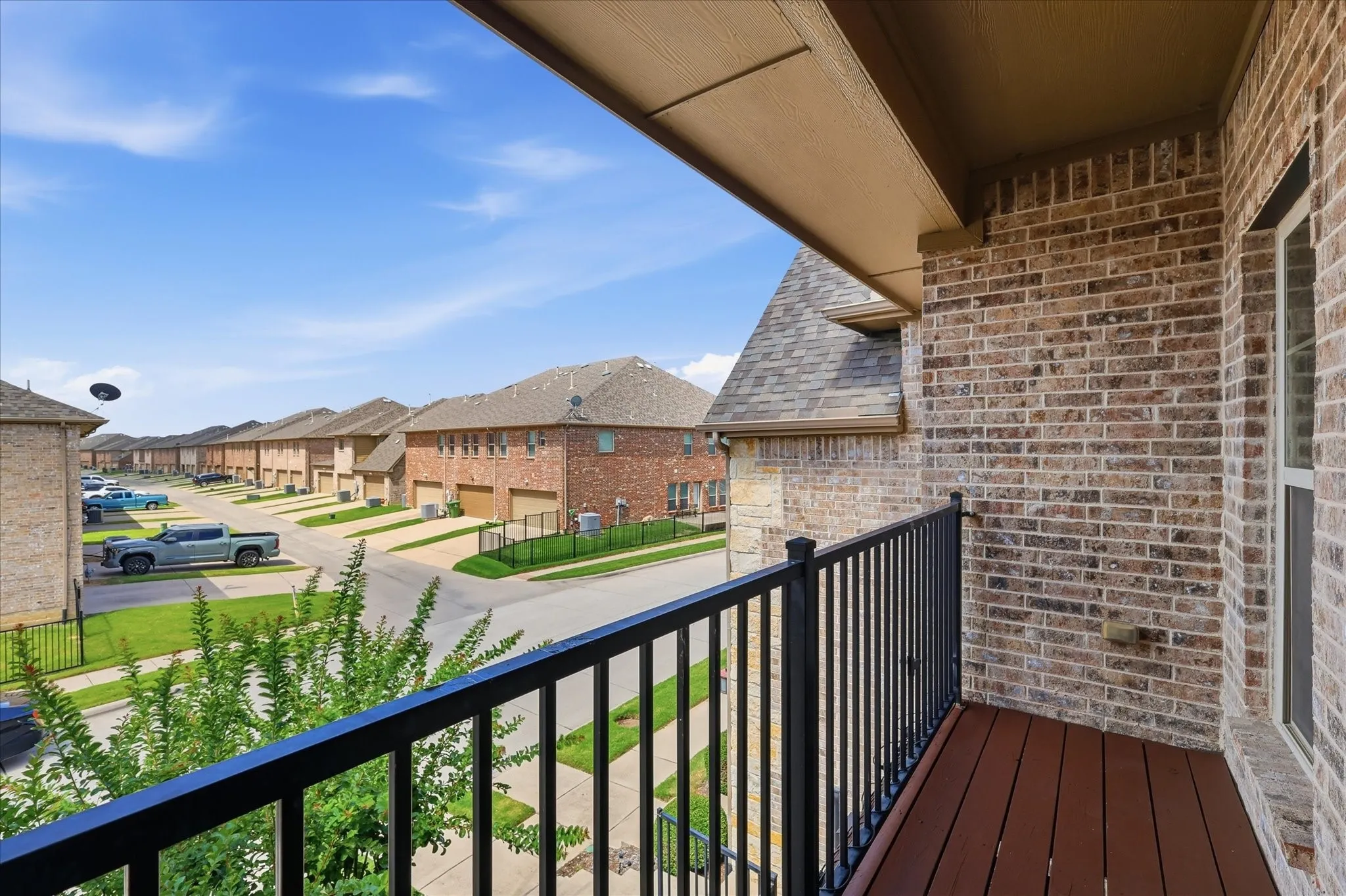 Private balcony with wood-finish flooring and black metal railing, featuring a brick exterior and a view of the residential streetscape