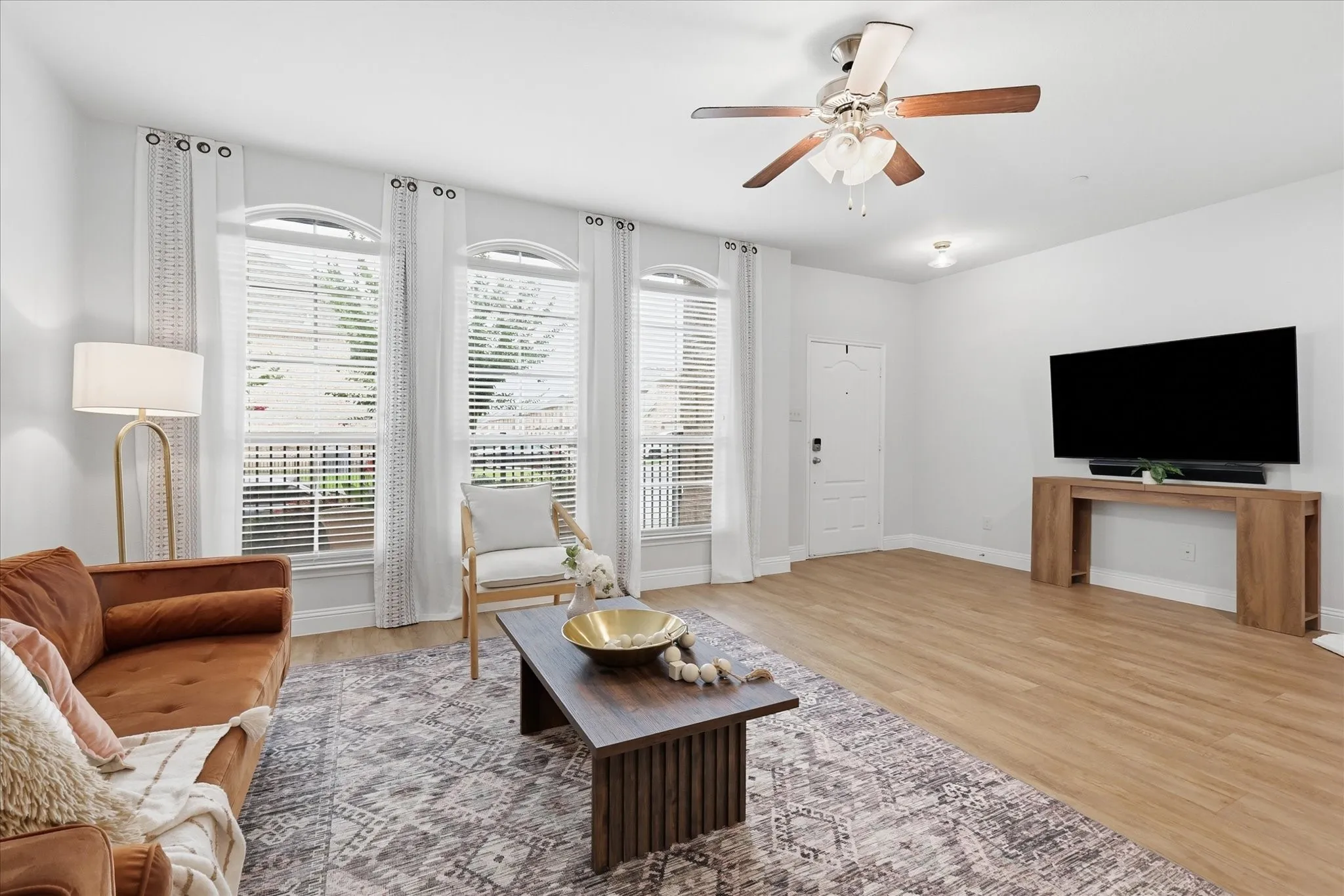 Spacious living area featuring wood-finish flooring, a ceiling fan with light fixture, and three arched windows with blinds
