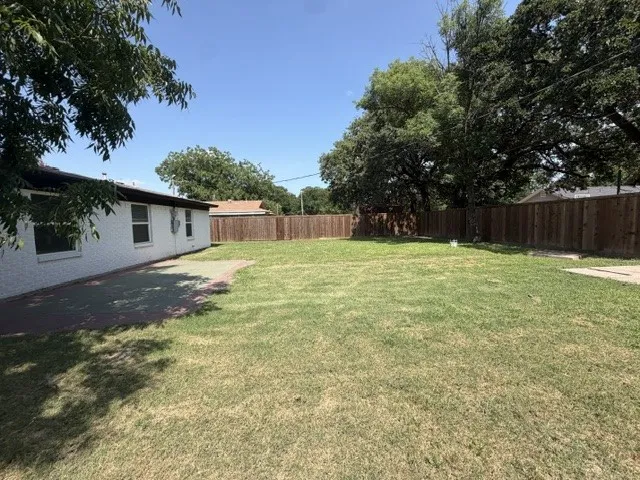Expansive backyard featuring a large grass area, wood privacy fencing, mature trees, a concrete patio, and a white brick exterior