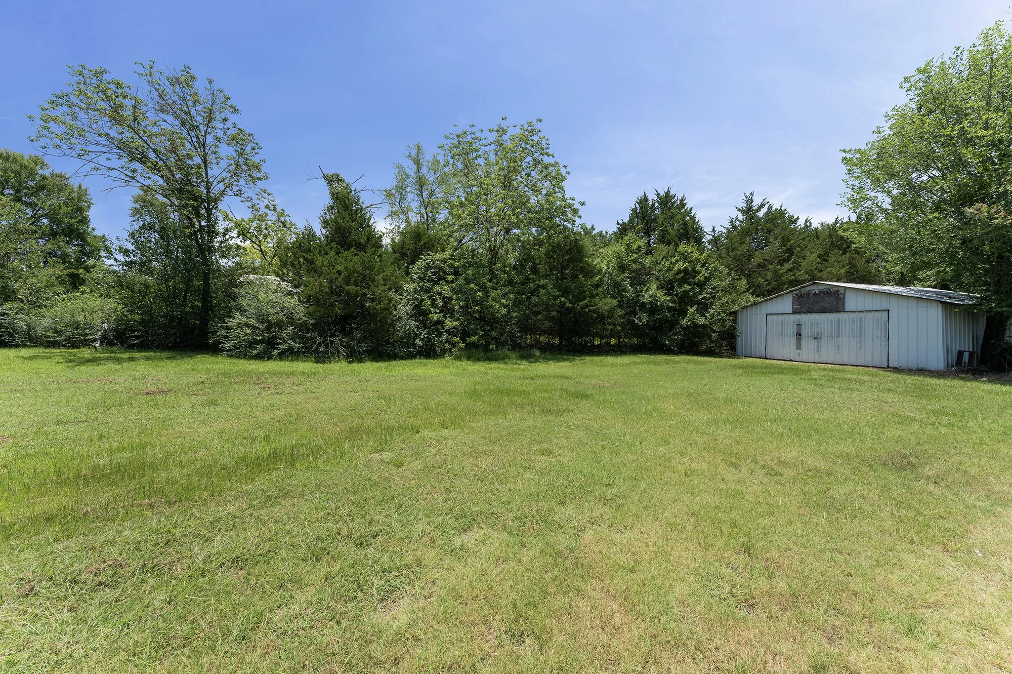 Expansive grassy area featuring a metal utility building with double doors and surrounding mature trees and shrubs