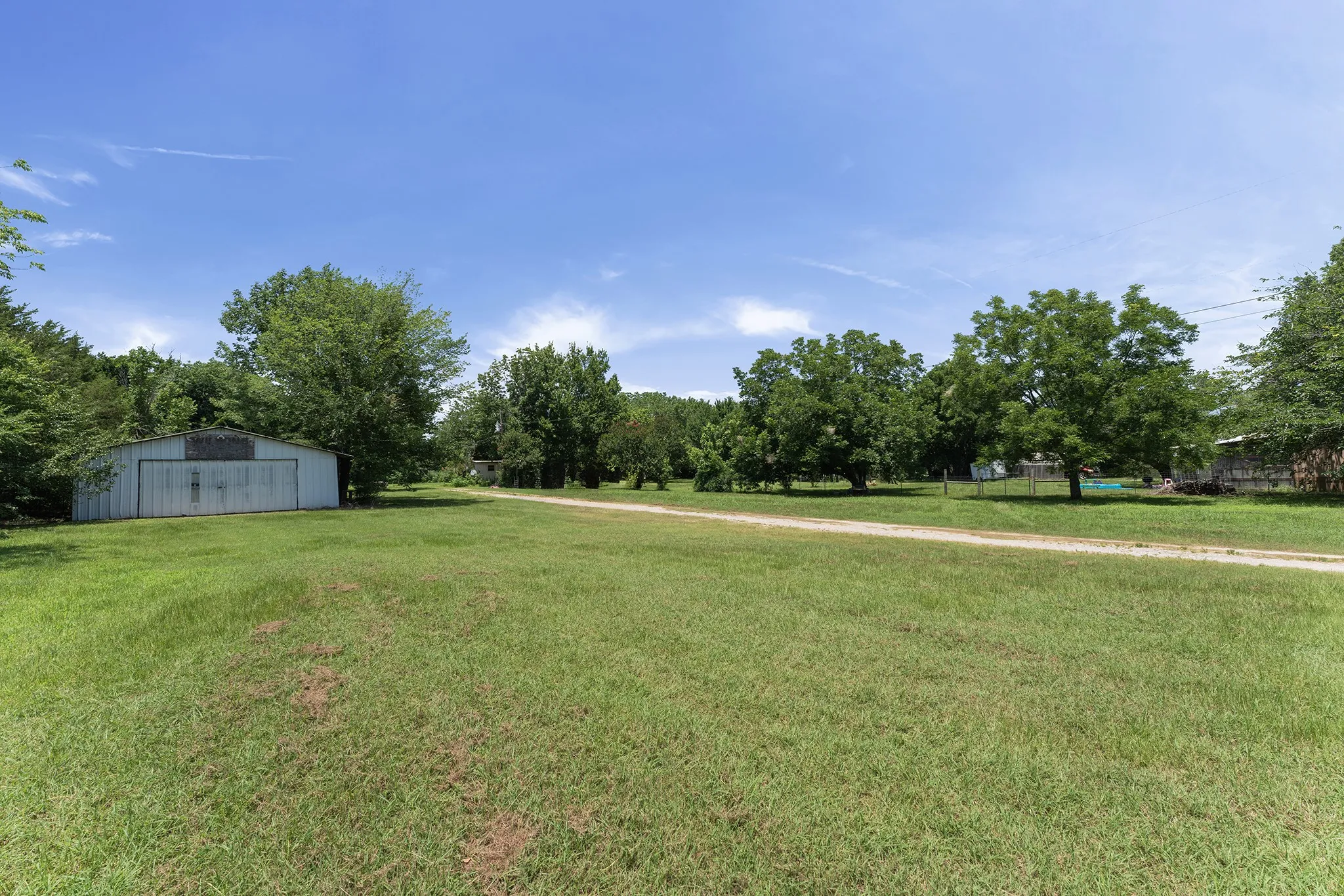 Expansive grass field featuring a long dirt driveway, mature trees, and a barn structure