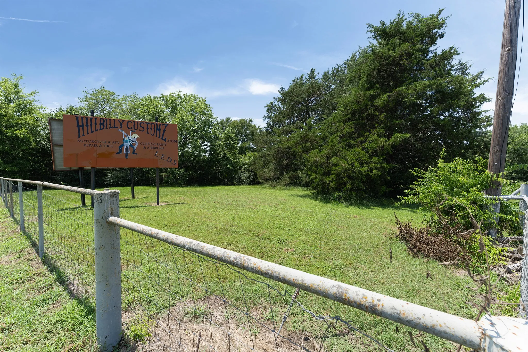 Expansive grassy lot with mature perimeter trees, featuring a chain-link fence with concrete posts