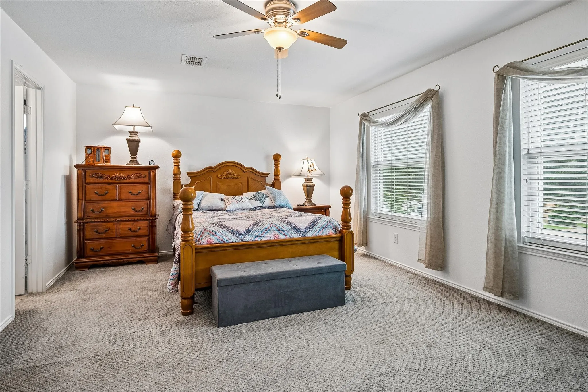 Tray ceiling, ceiling fan with integrated lighting, two windows with blinds, and neutral carpeting