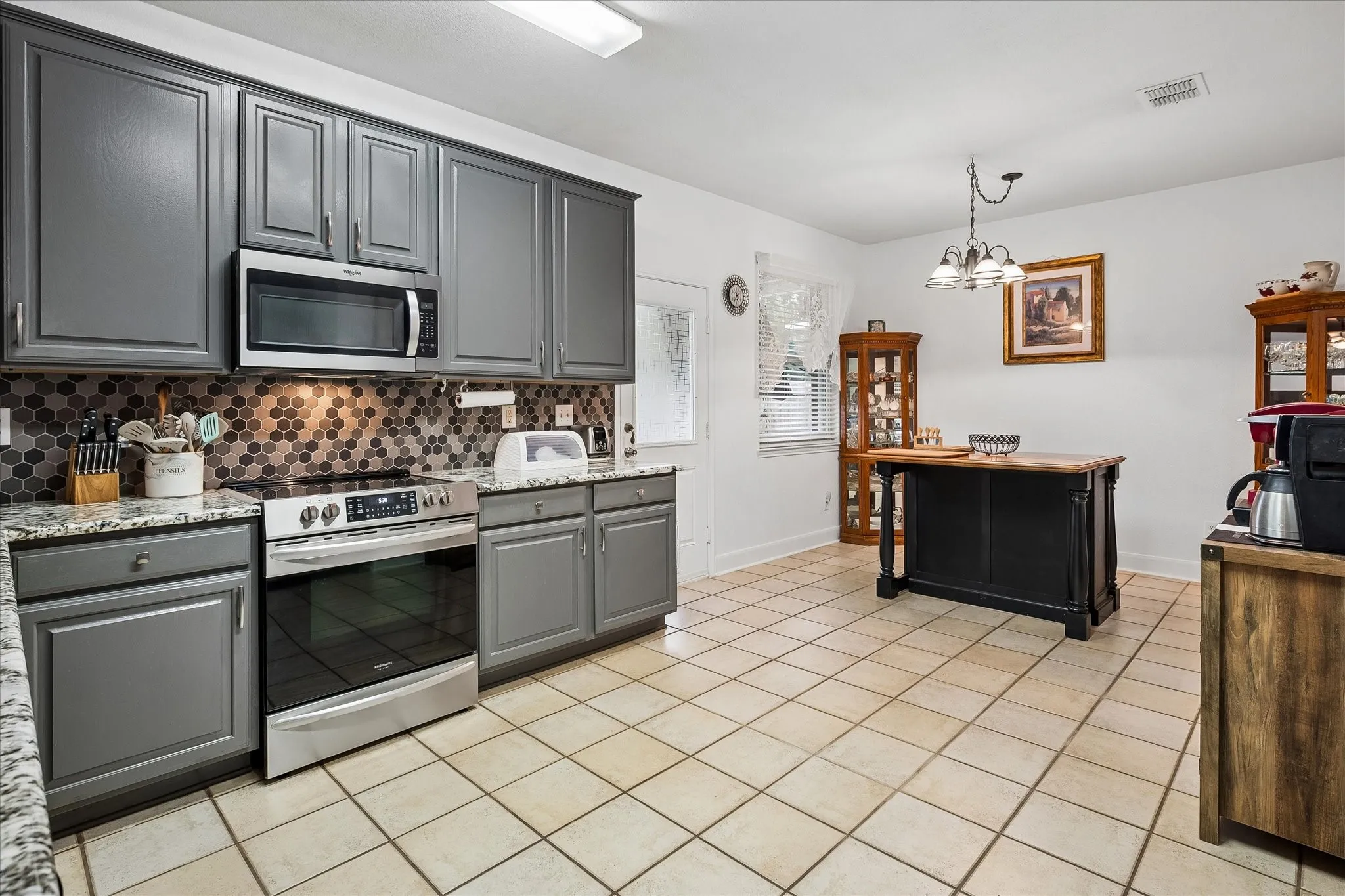 Gray cabinetry with stainless steel hardware, hexagonal tile backsplash, stainless steel range and microwave, light-toned tile flooring, and a central freestanding island