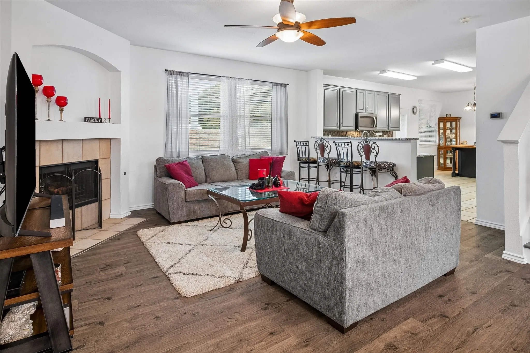 Wood-finish flooring, tiled fireplace with hearth, ceiling fan, and window with blinds