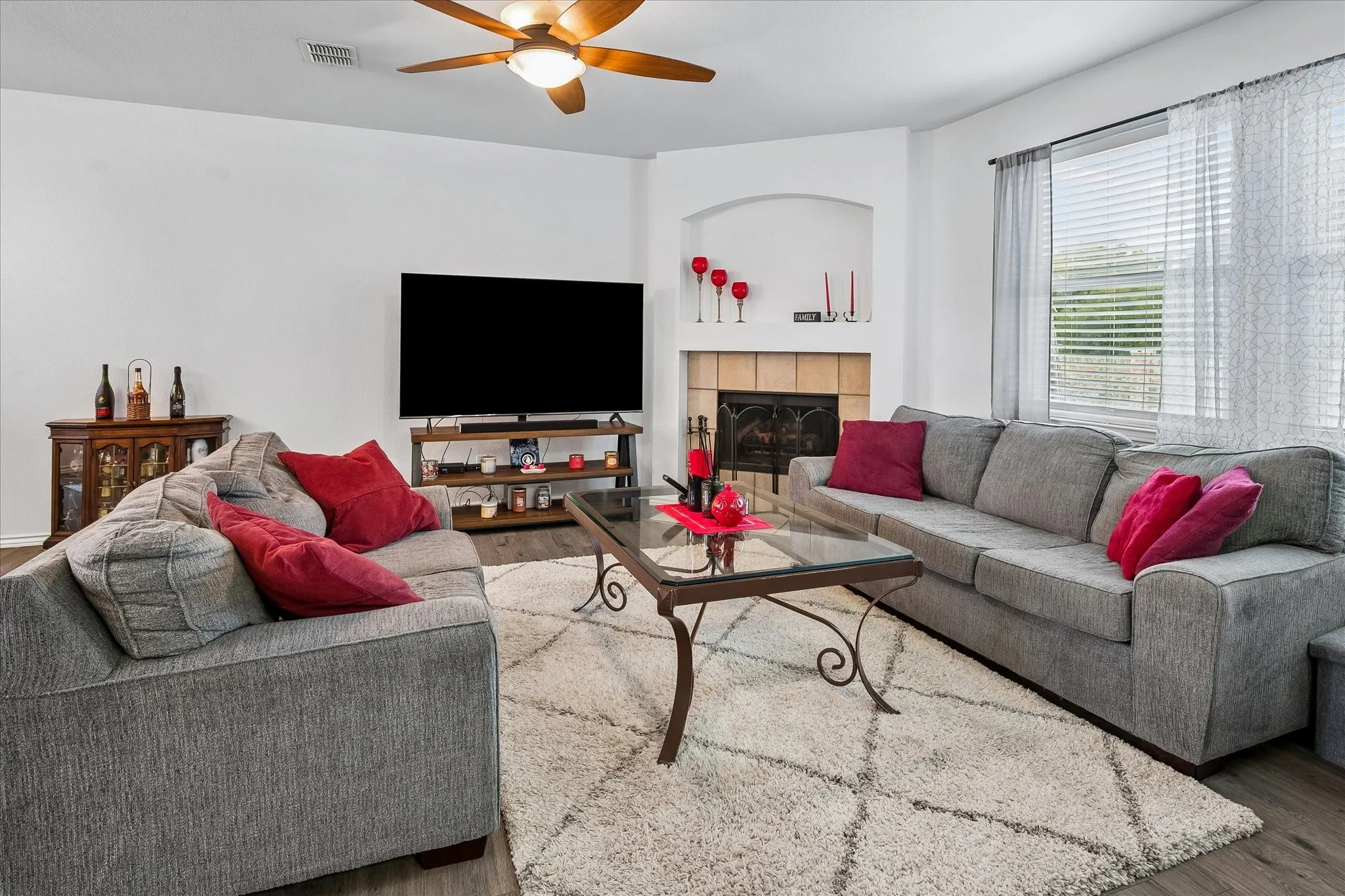 Living area featuring wood-finish flooring, a ceiling fan, a fireplace with a tiled surround, and a window with blinds