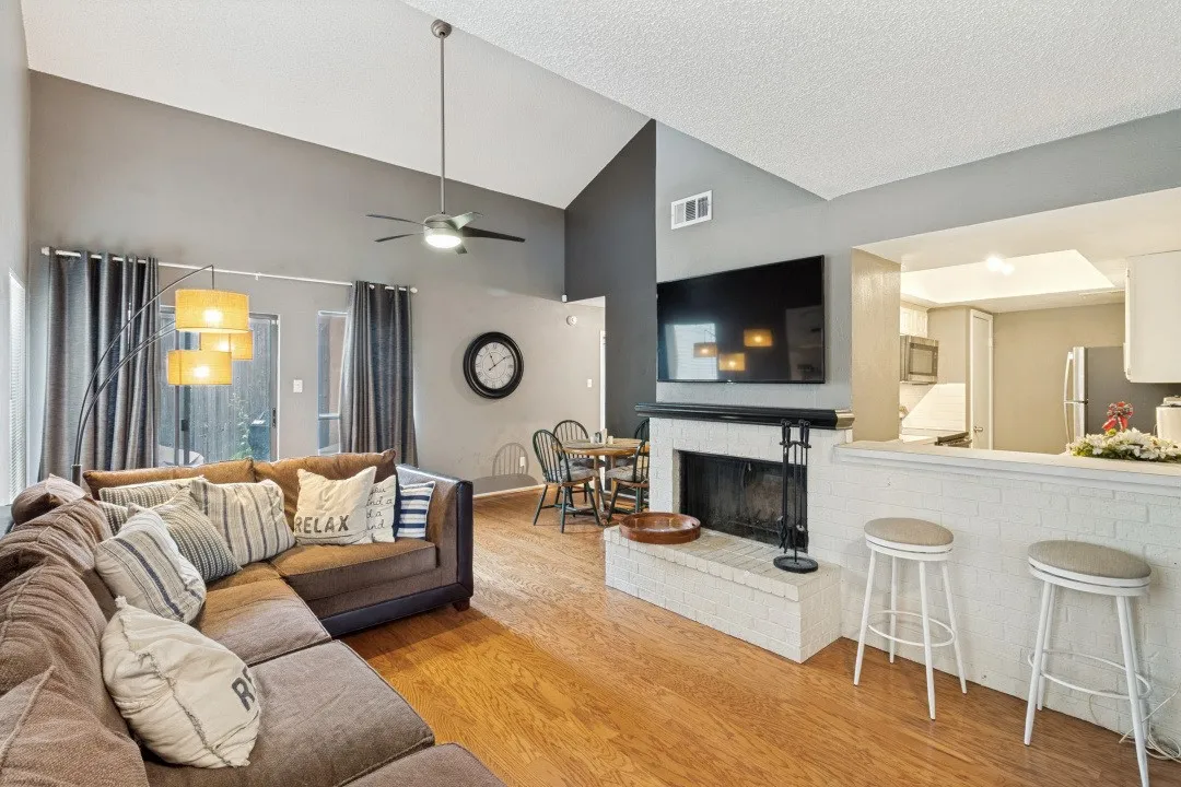Vaulted ceiling with a ceiling fan, fireplace with a white brick hearth and black mantel, wood-finish flooring, and a kitchen pass-through with a white brick half-wall