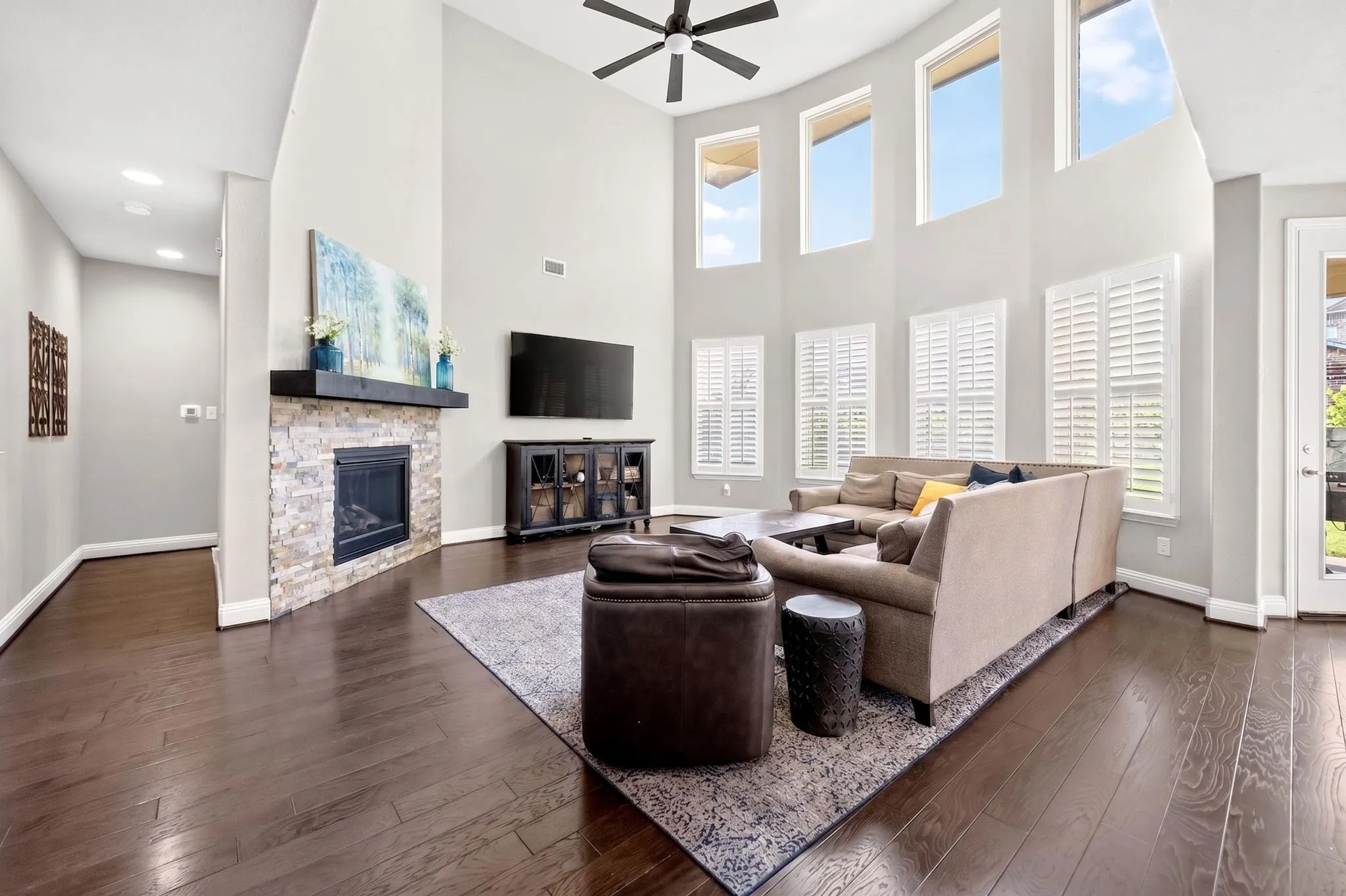 Two-story living area featuring dark wood-finish flooring, a stone-clad fireplace with a dark mantel, and multiple windows with plantation shutters