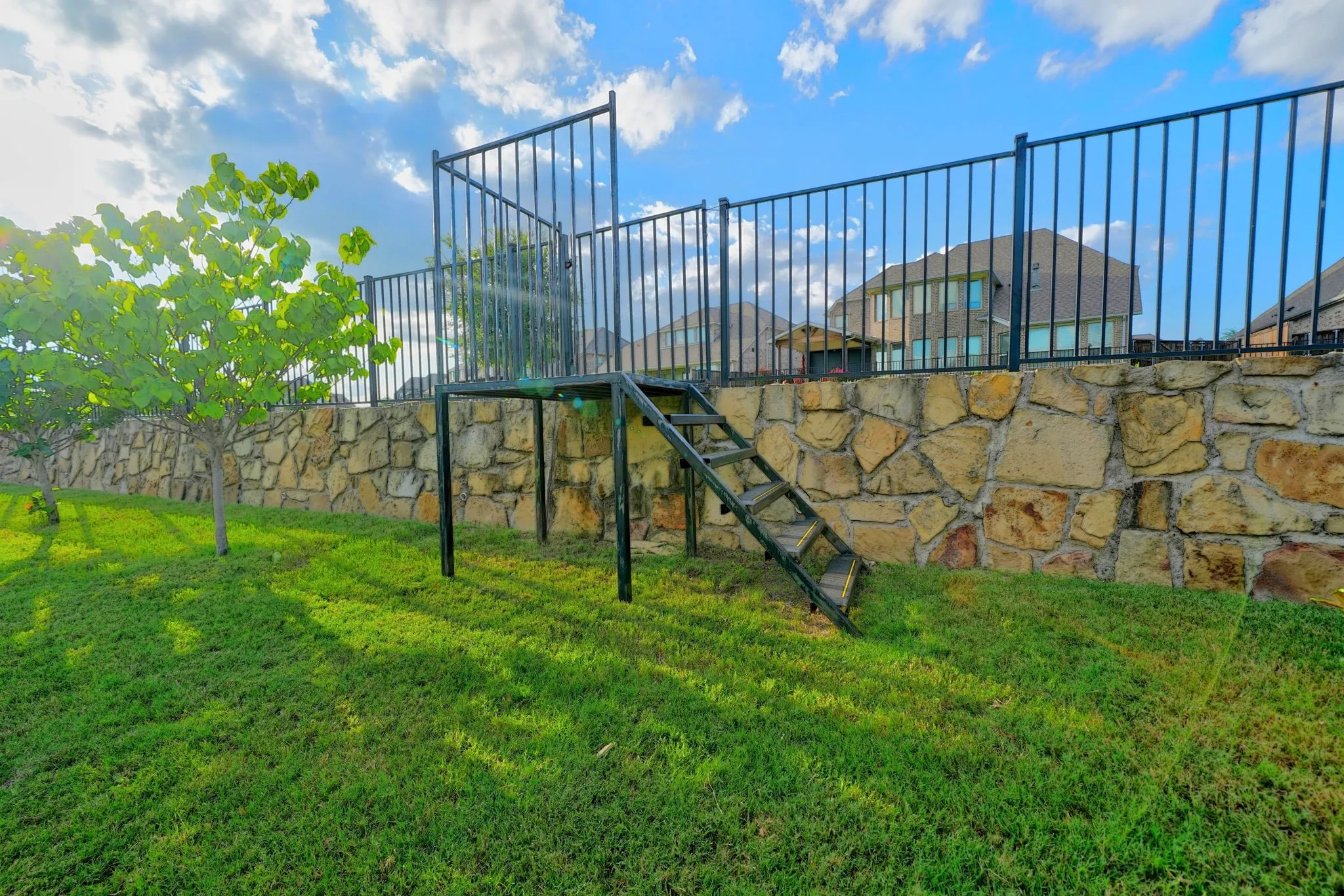 Expansive backyard featuring a manicured lawn, stone retaining wall, and black wrought iron fencing
