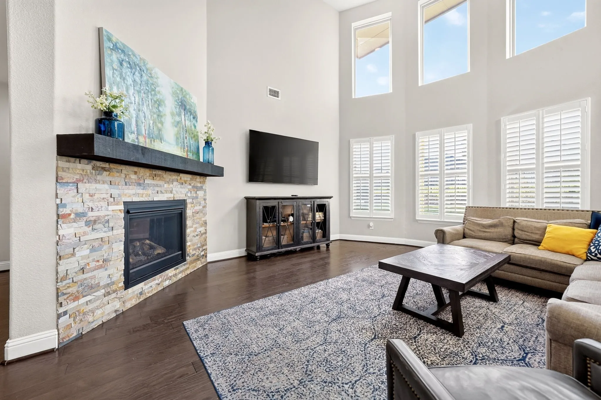 Living area featuring a stone-faced fireplace with a dark wood mantel, high ceilings, dark wood-finish flooring, and multiple windows with white plantation shutters