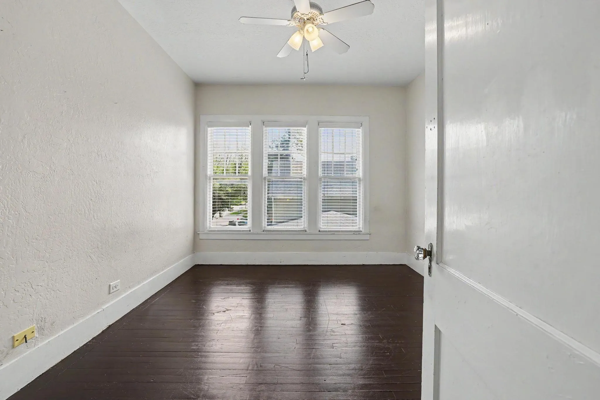 Room featuring dark wood-finish flooring, white baseboards, and a three-panel window with horizontal blinds