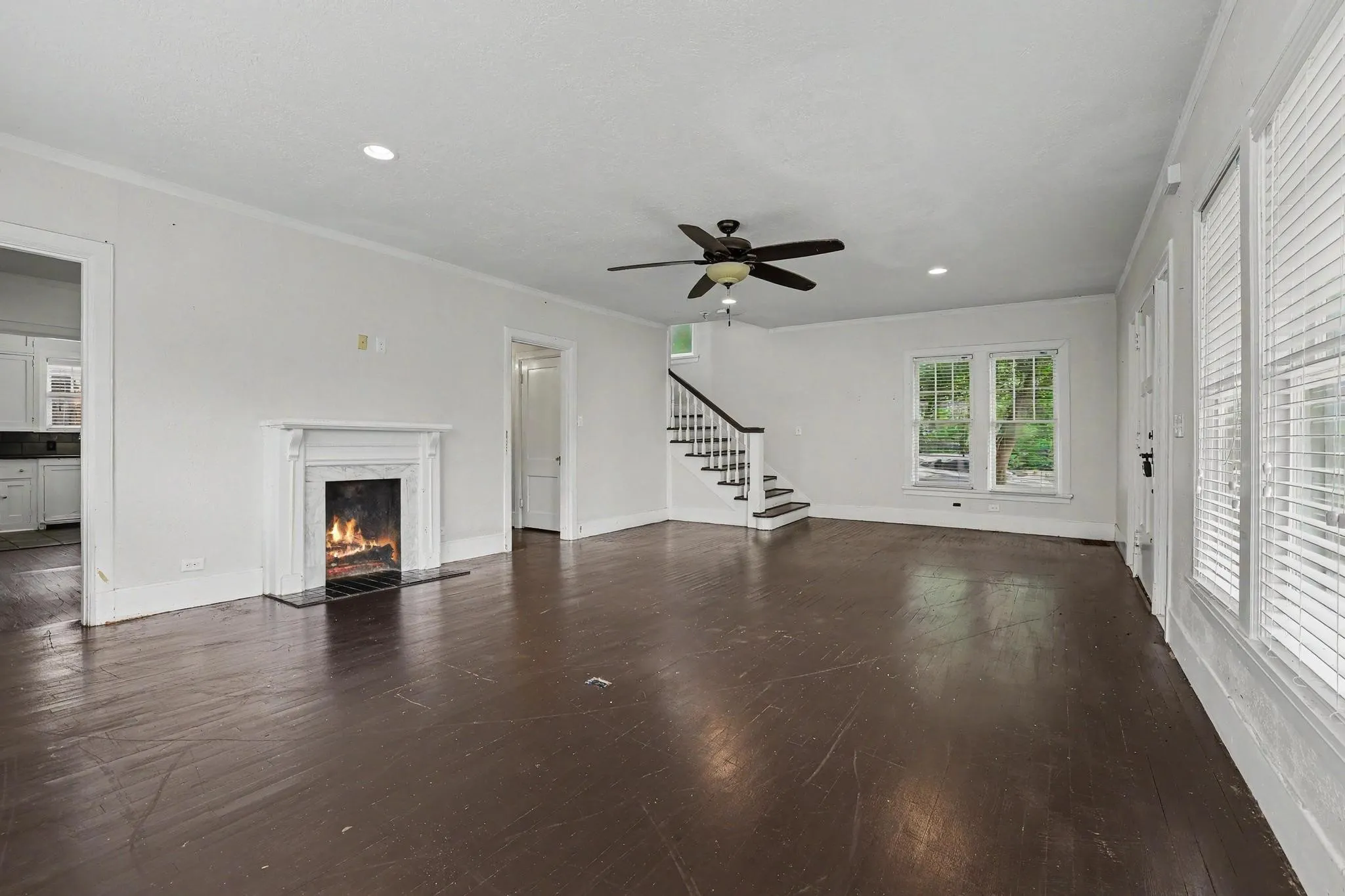 Spacious living area featuring dark wood-finish flooring, a white mantel fireplace with a dark stone hearth, a ceiling fan, recessed lighting, and multiple windows with blinds