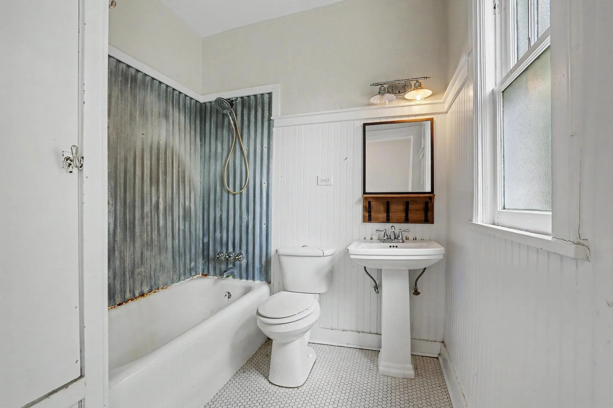 Bathroom featuring a pedestal sink, a bathtub with corrugated metal surround, white beadboard paneling, mosaic tile flooring, and a window