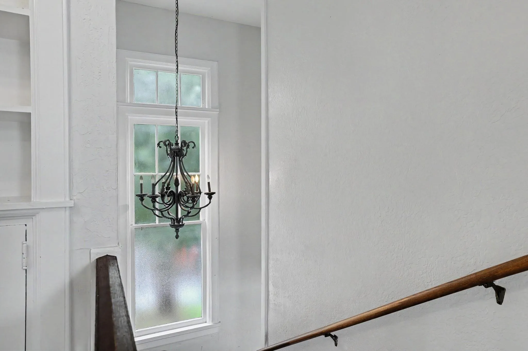 Decorative chandelier with multiple candelabra bulbs, positioned before a tall window featuring a transom and white trim
