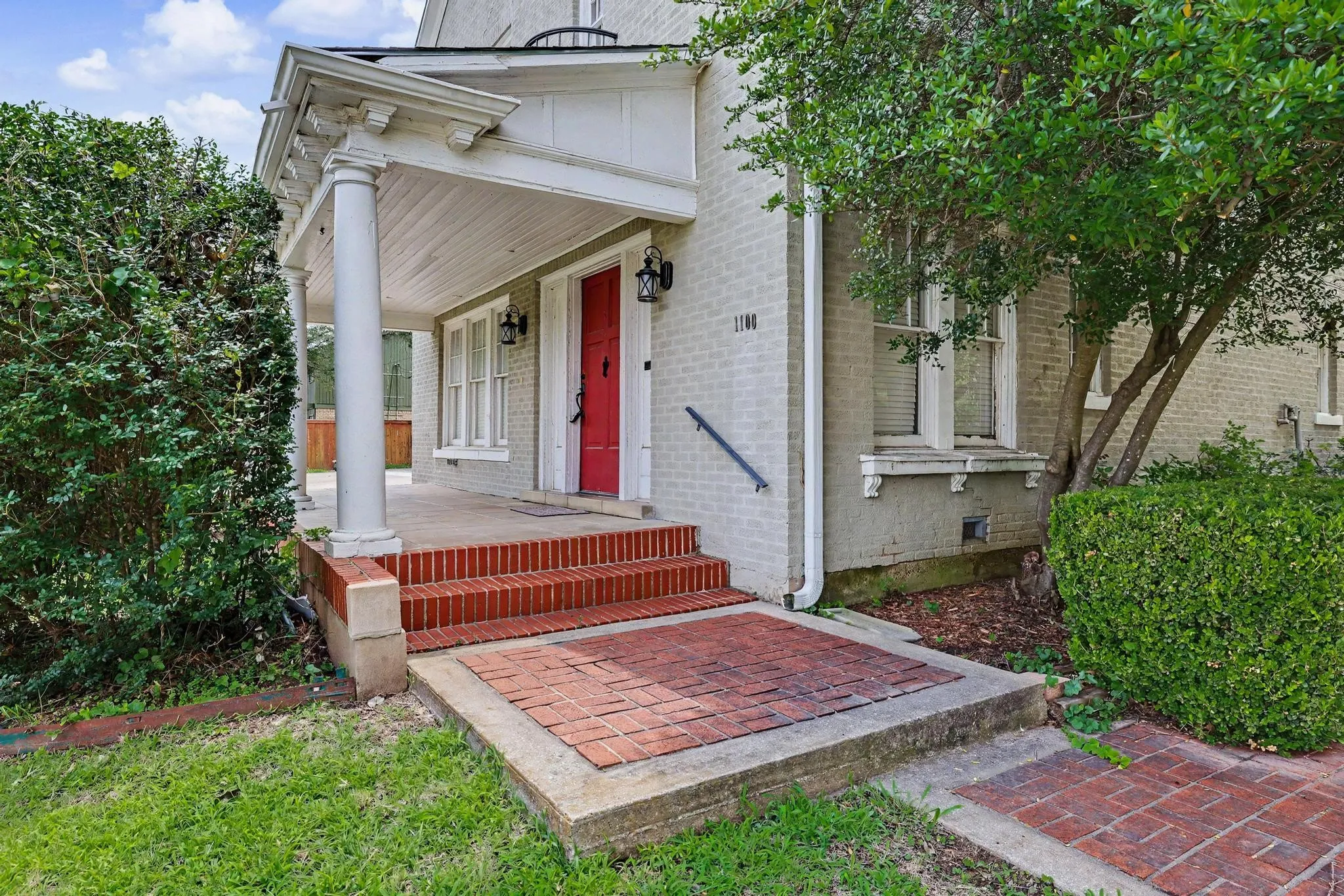 Brick exterior featuring a covered portico with decorative columns, a red entry door, brick steps, and established landscaping