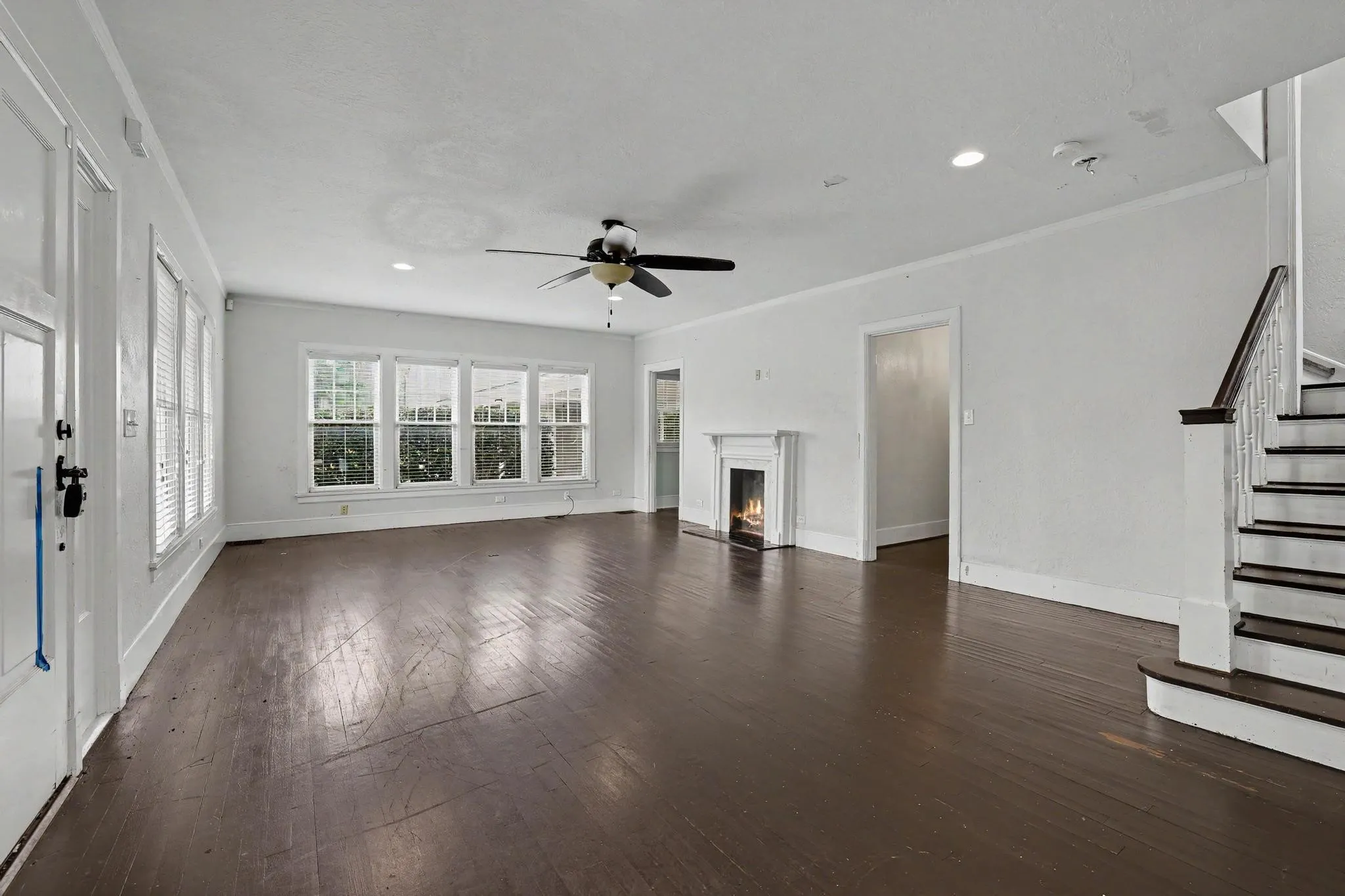 Spacious living area featuring wood-finish flooring, a traditional fireplace with a white mantel, multiple windows, a ceiling fan, and a staircase with white risers and dark treads