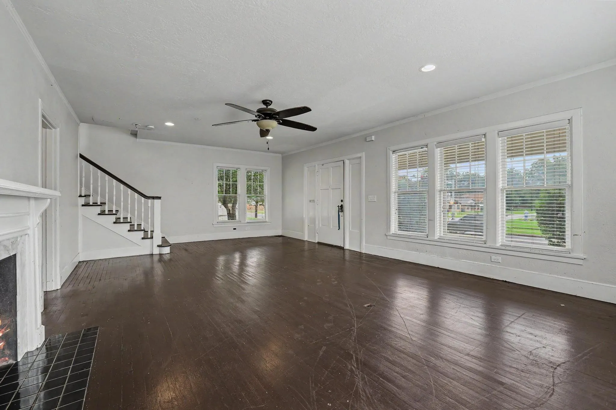 Spacious living area featuring dark wood-finish flooring, a white mantel fireplace with black tile surround, a staircase with white spindles and dark handrail, a ceiling fan, and multiple windows with blinds