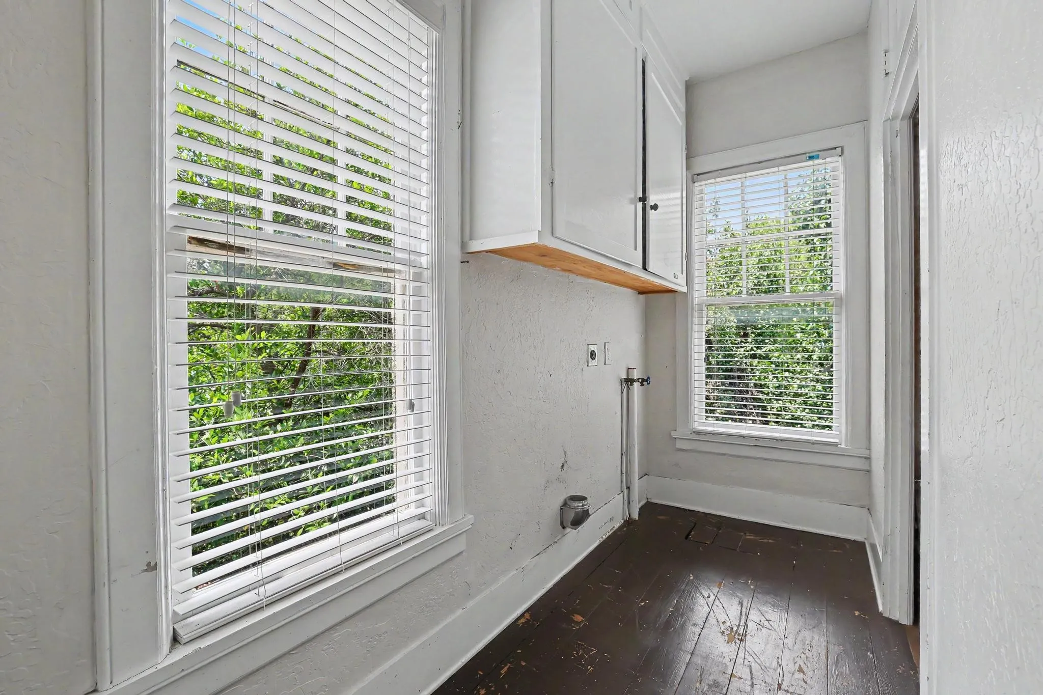 Dedicated utility area featuring two windows with white blinds, overhead white cabinetry with a wood-finish shelf, and dark wood-finish flooring