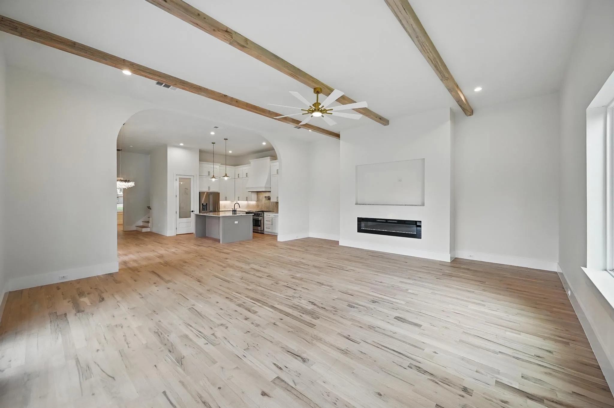 Spacious living area featuring light wood-finish flooring, exposed ceiling beams, recessed lighting, and a modern electric fireplace