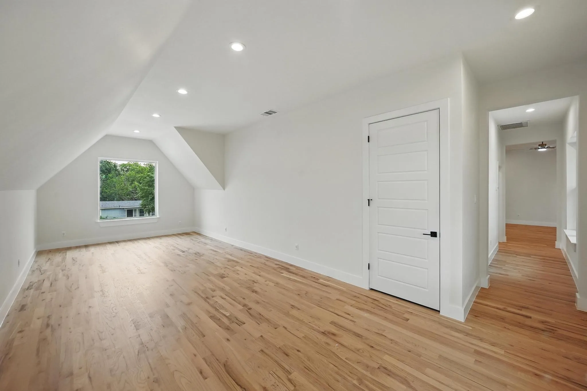 Spacious room featuring hardwood flooring, recessed lighting, a window with exterior views, and a five-panel interior door with matte black hardware