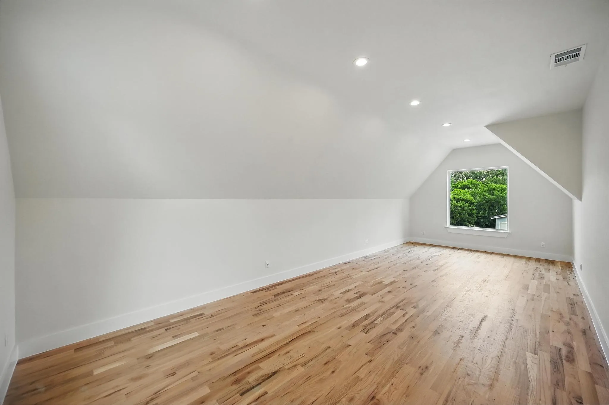 Spacious room featuring sloped ceilings, recessed lighting, hardwood flooring, and a window overlooking lush greenery
