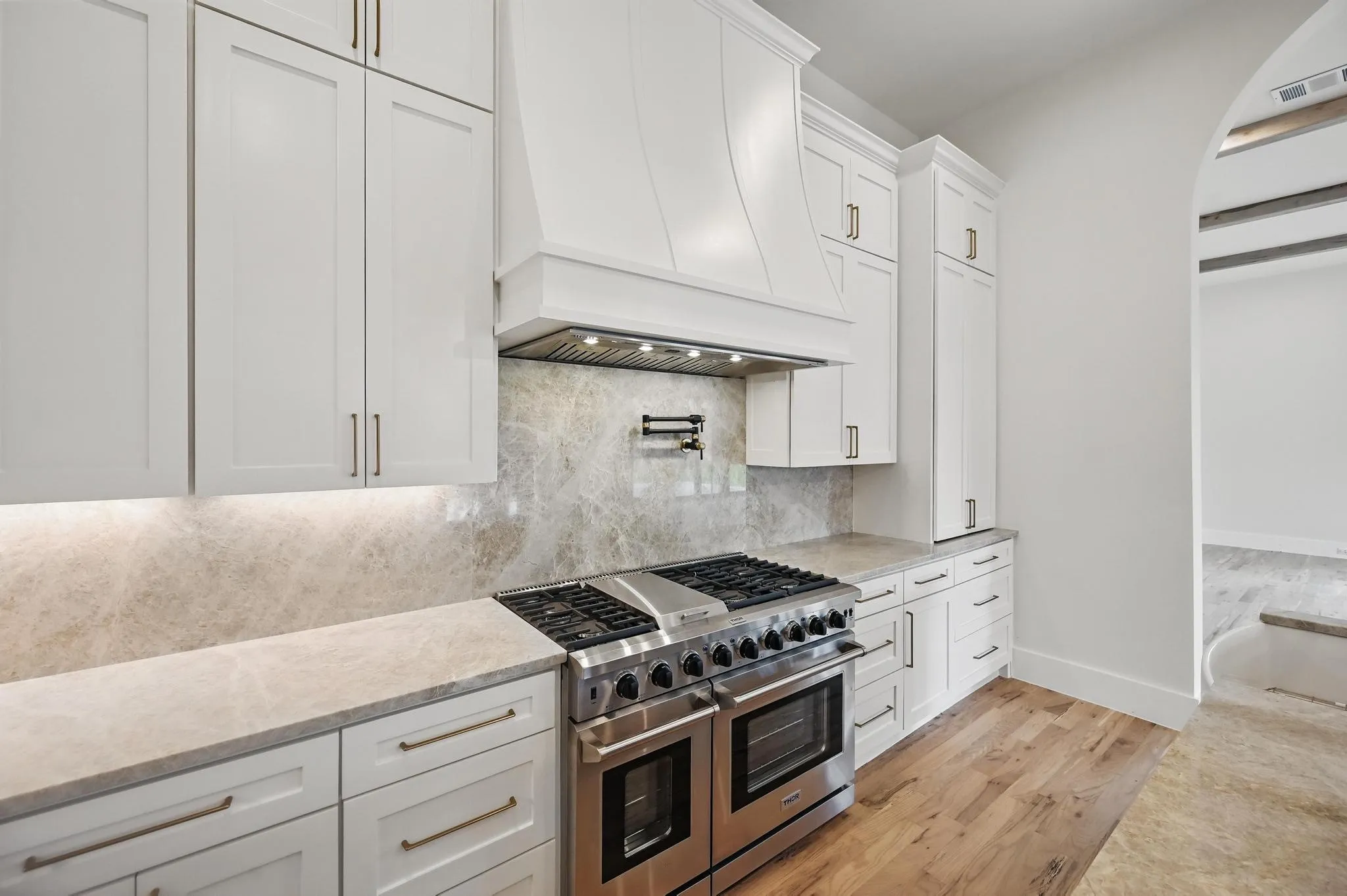 Gourmet kitchen featuring a stainless steel gas range with double ovens, a built-in range hood, white shaker-style cabinetry with gold-tone hardware, light-toned stone countertops, and a full-height stone backsplash