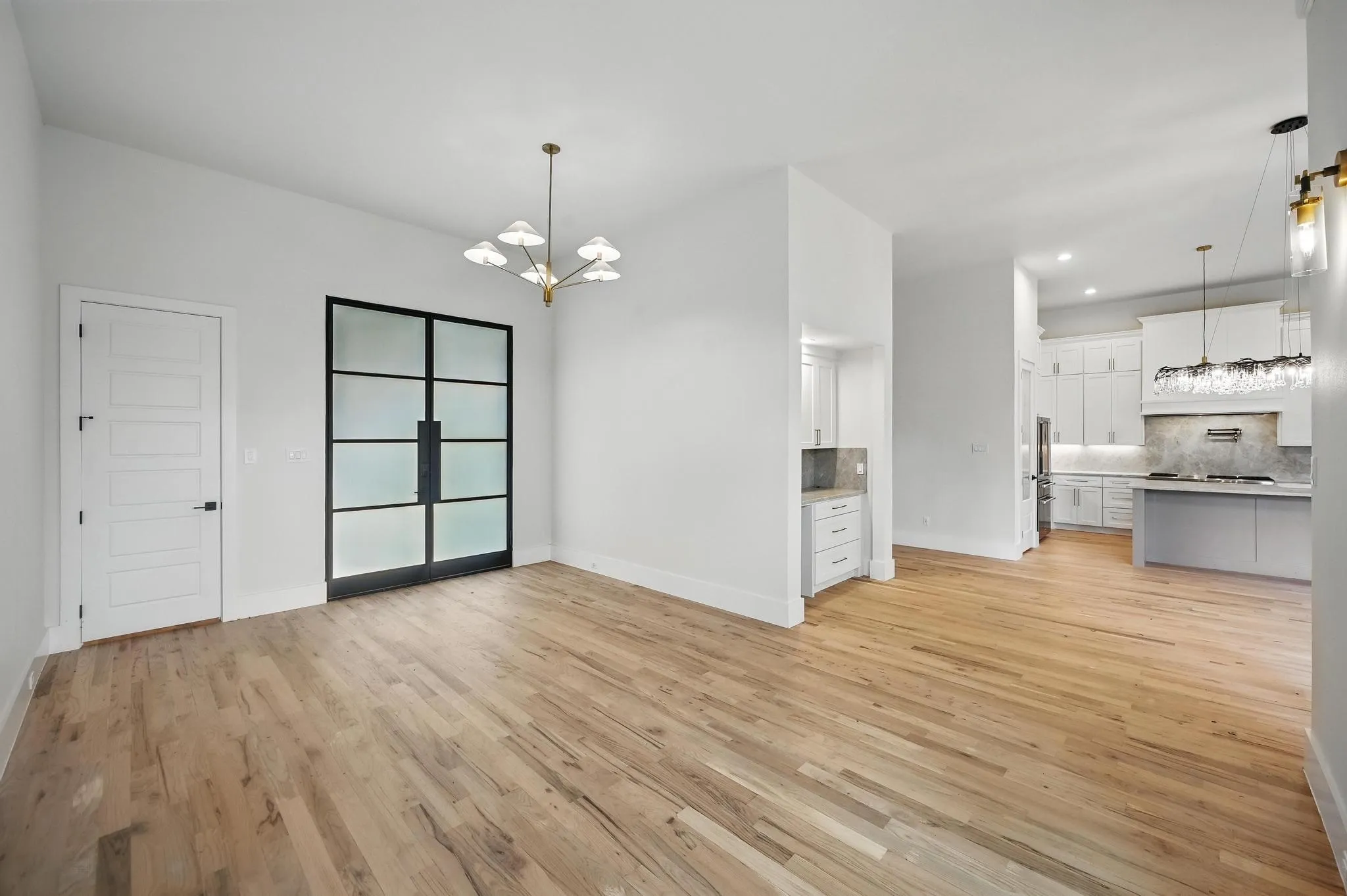 Expansive room featuring light wood-finish flooring, a modern chandelier, and a black-framed double entry door with frosted glass panels