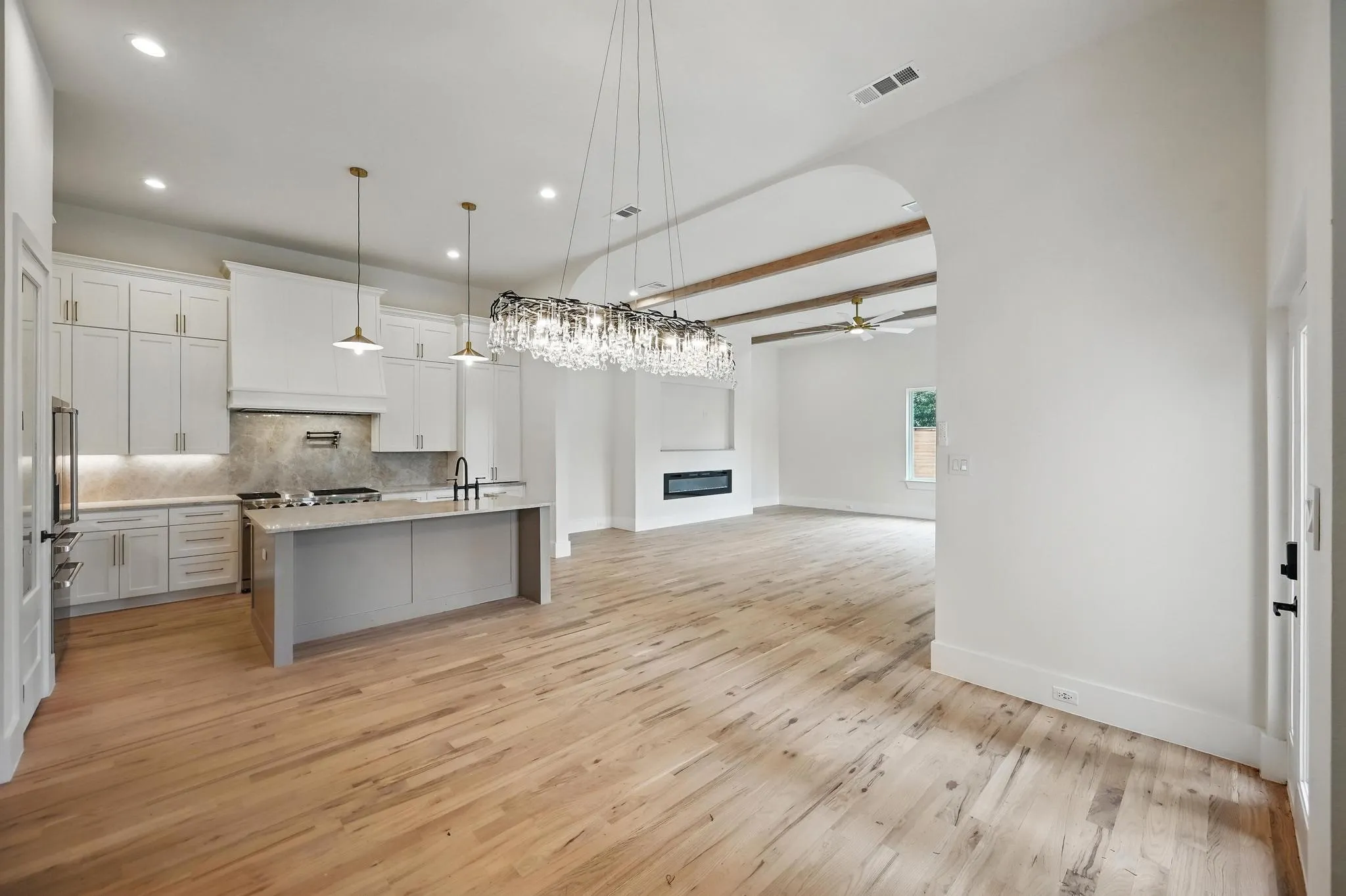 Open-concept kitchen and living area featuring wood-finish flooring, a kitchen island with pendant lighting, white cabinetry, a built-in electric fireplace, and exposed ceiling beams