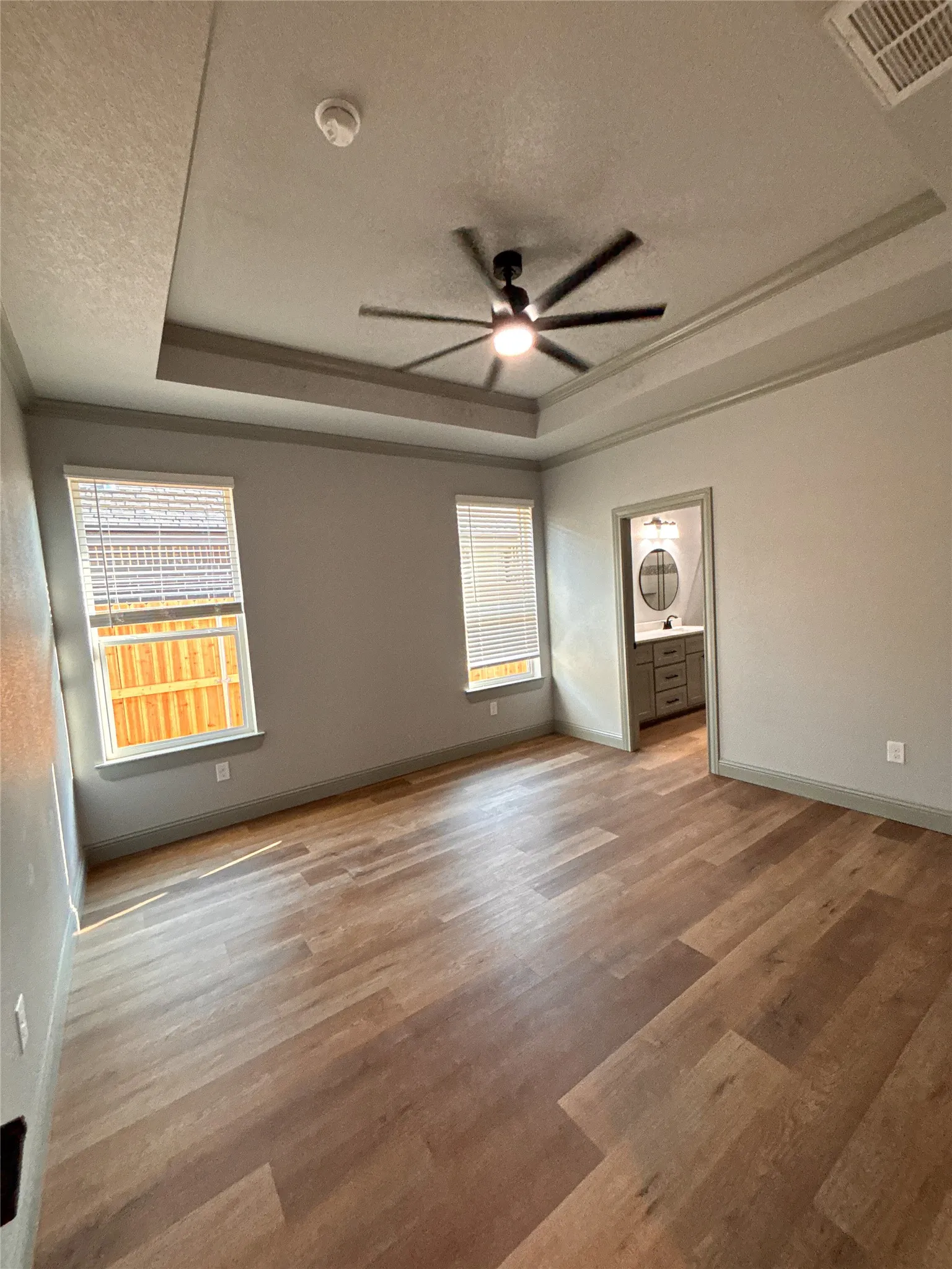 Spacious room featuring wood-finish flooring, two windows with blinds, and a tray ceiling with a ceiling fan