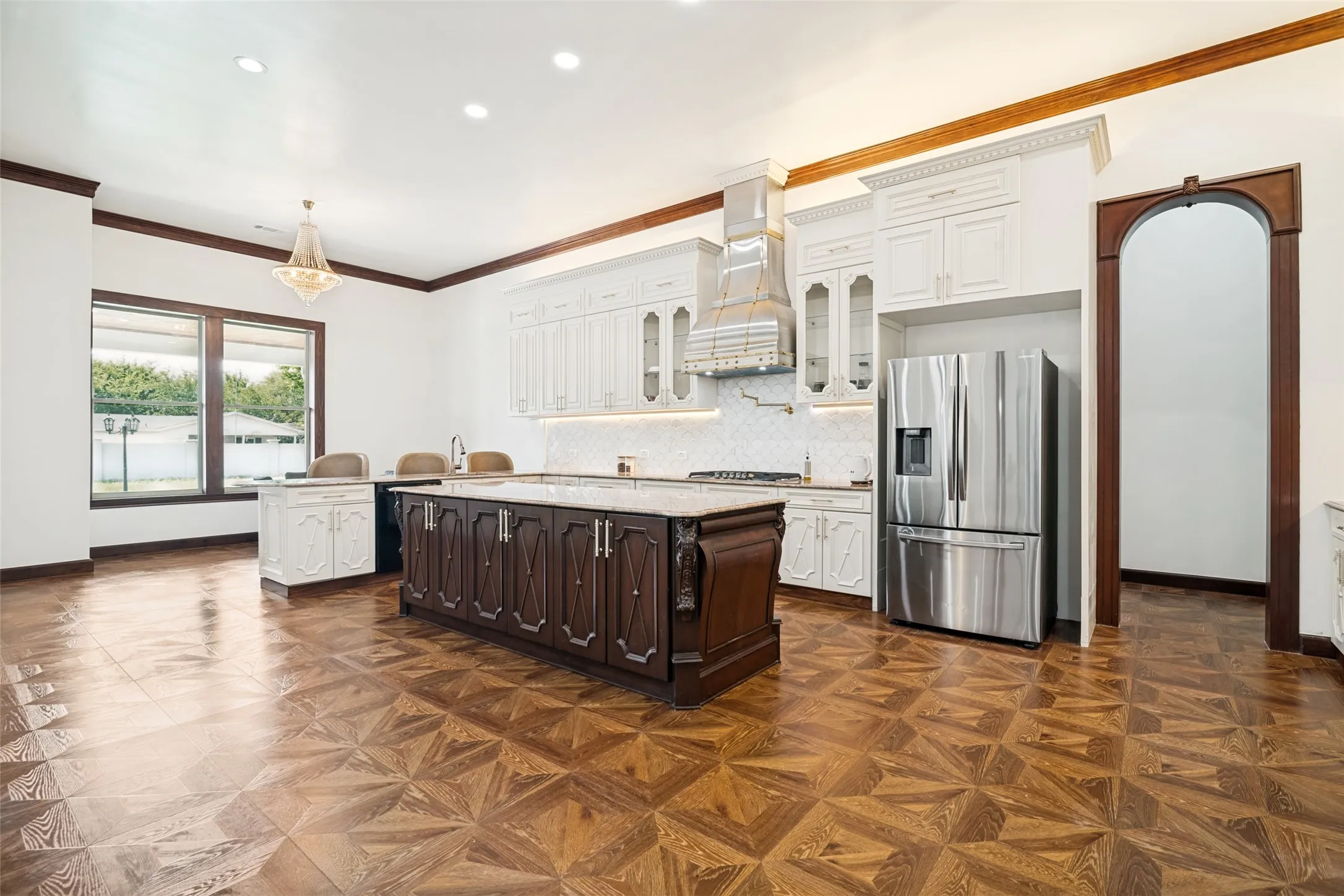 Expansive kitchen featuring a large central island with a stone countertop, white upper cabinetry, stainless steel appliances, and wood-finish flooring with a geometric pattern