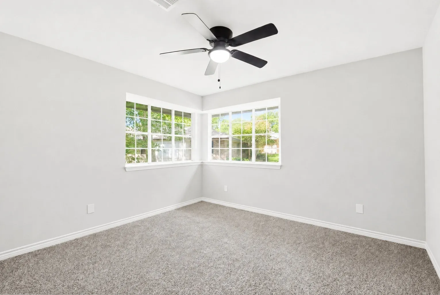 Bright room featuring corner windows, gray carpet, white baseboards, and a black ceiling fan with integrated lighting